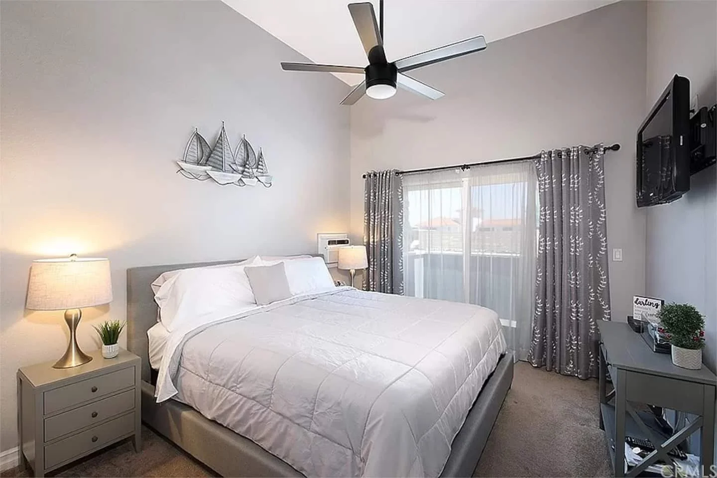 A bedroom with a large bed with white bedding, beige pillows, and a gray headboard. There are two lamps on nightstands on either side of the bed. A metal wall art of sailboats hangs above the bed. A sliding glass door with sheer curtains and patterned drapes leads outside. A wall-mounted TV is on the right side, and a gray desk with a potted plant and decorative items is below it. A ceiling fan with lights is on the ceiling.