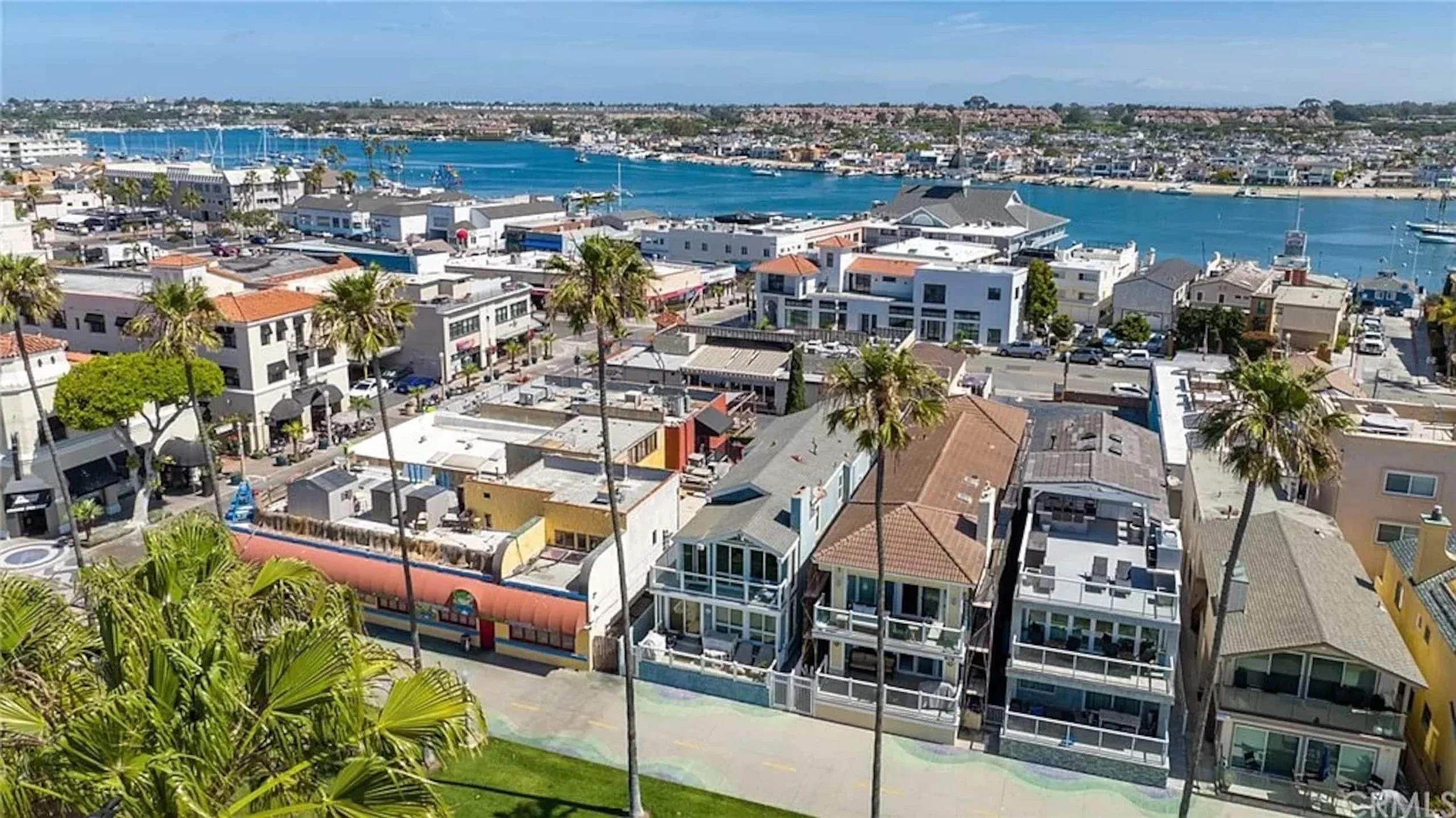 Aerial view over the Balboa Peninsula, with the beachfront boardwalk, the row of oceanfront homes, and Newport Harbor stretching out beyond.
