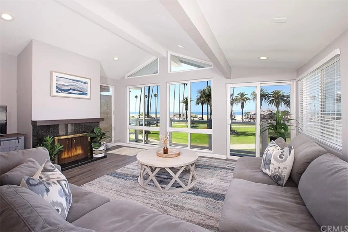 Bright living room with large windows and view of palm trees and ocean, featuring a fireplace, beige sofas, round coffee table, and houseplants.