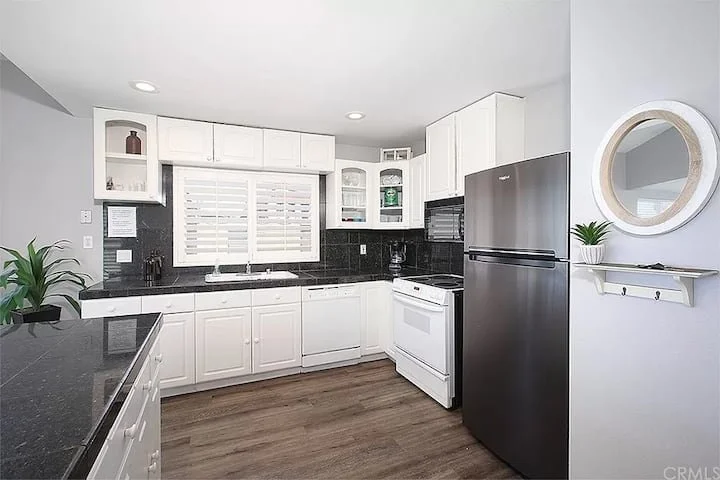 Kitchen with white cabinets, black countertops, a white stove, stainless steel refrigerator, and black backsplash. There is a window with shutters, a round mirror on the wall, and a green plant on a white shelf.