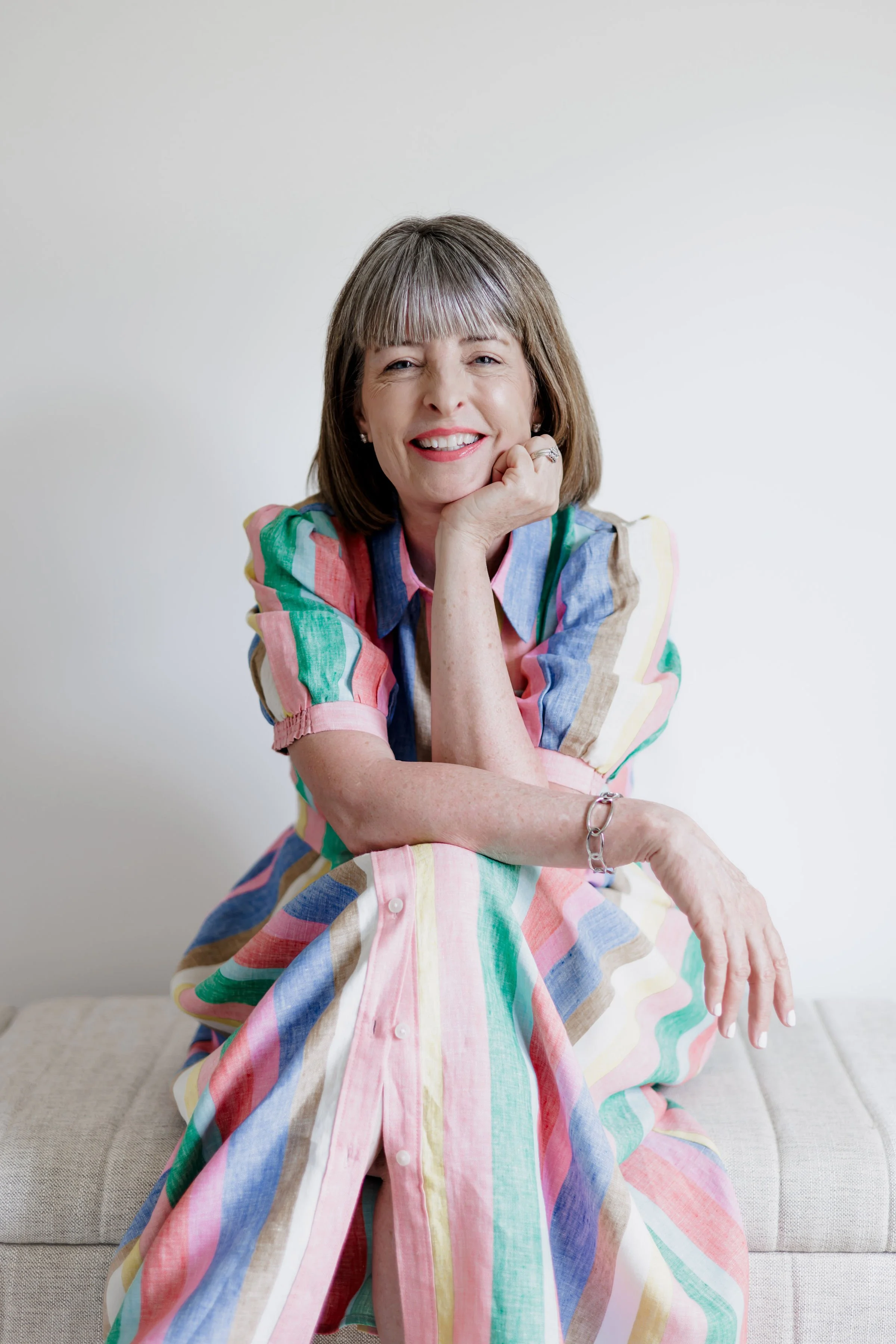 A smiling middle-aged woman with short brown hair and bangs, wearing a colorful, striped dress, resting her chin on her hand, sitting on a beige couch against a plain light gray wall.