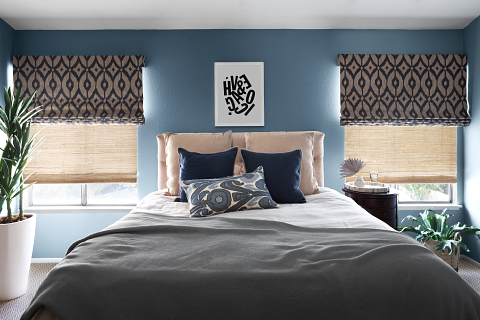 Bedroom with a bed, navy and patterned pillows, framed artwork, two windows with patterned blinds, side table, and plants.
