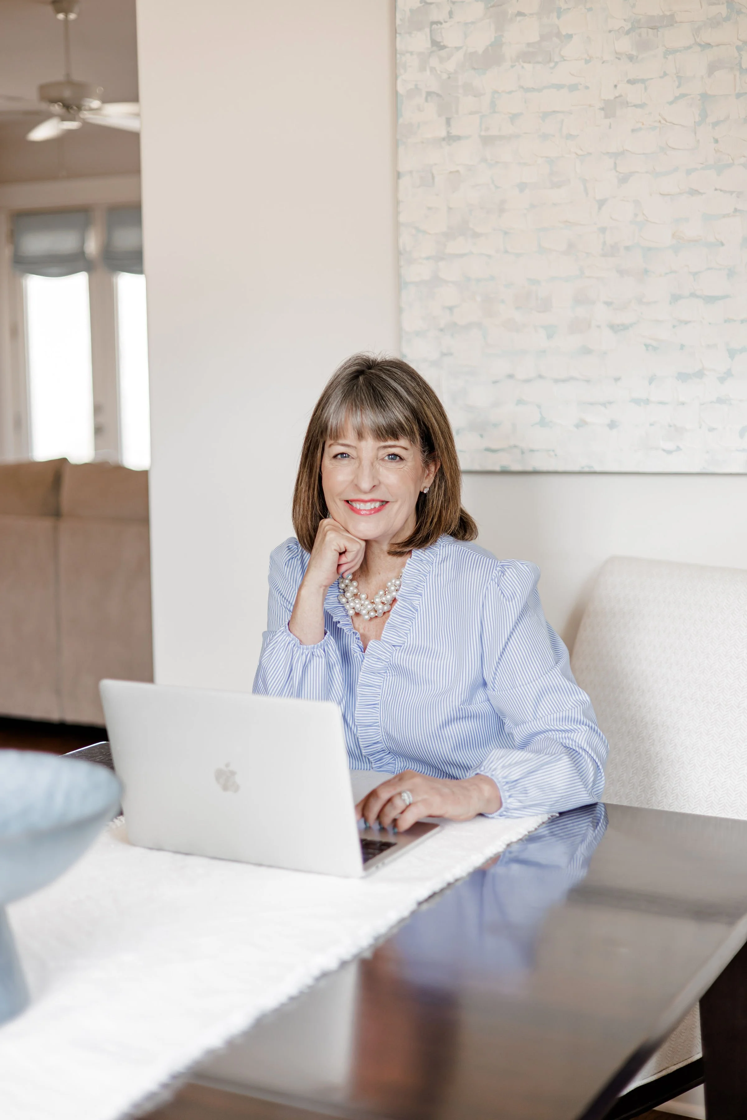 A middle-aged woman with brown hair and a pearl necklace sitting at a dining table with a silver MacBook laptop, smiling and resting her chin on her hand, in a bright dining room with abstract art on the wall and a beige couch in the background.
