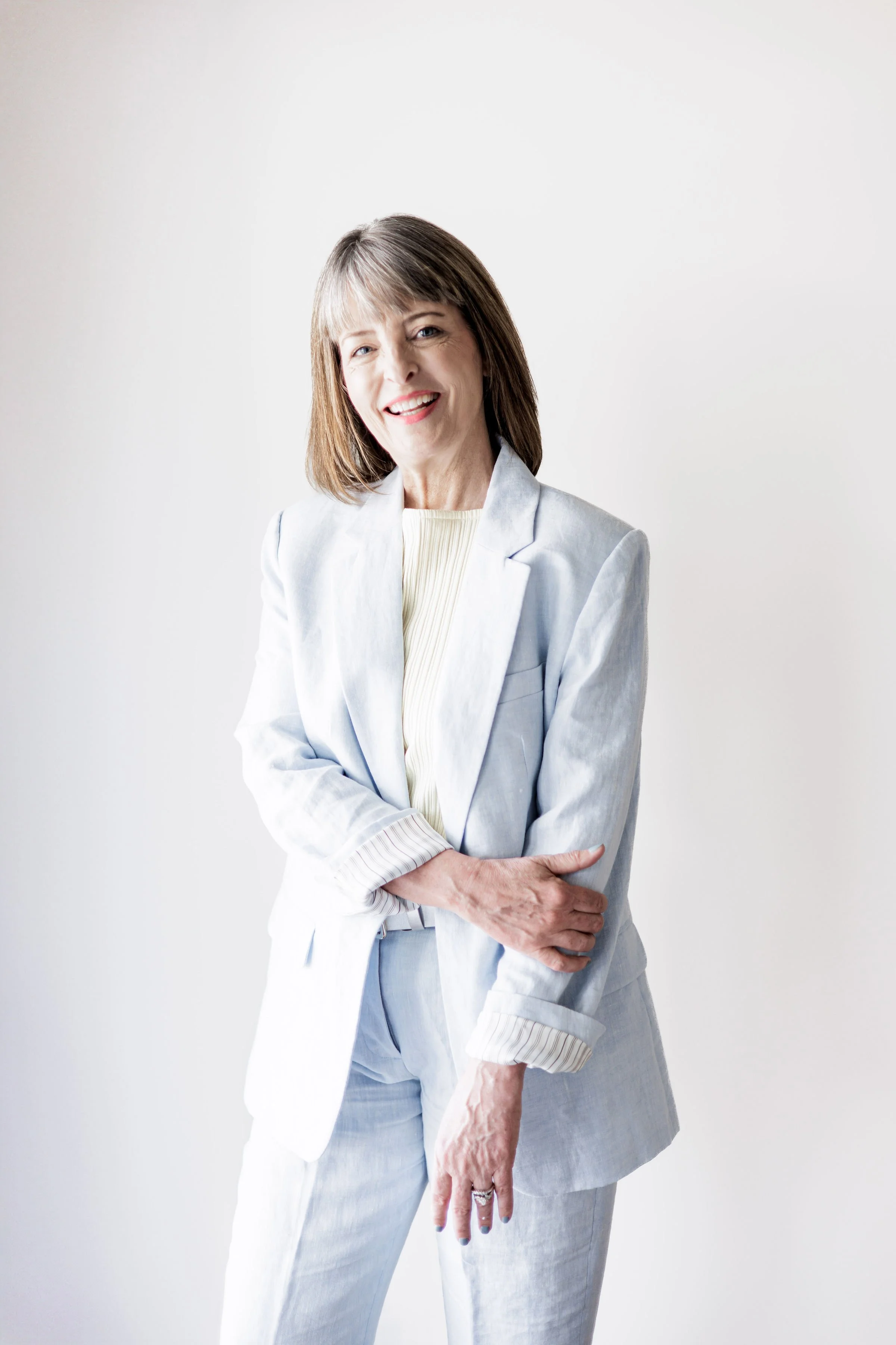 Portrait of a smiling middle-aged woman with shoulder-length brown hair, wearing a light blue blazer over a white top and light blue pants, standing against a plain white wall.