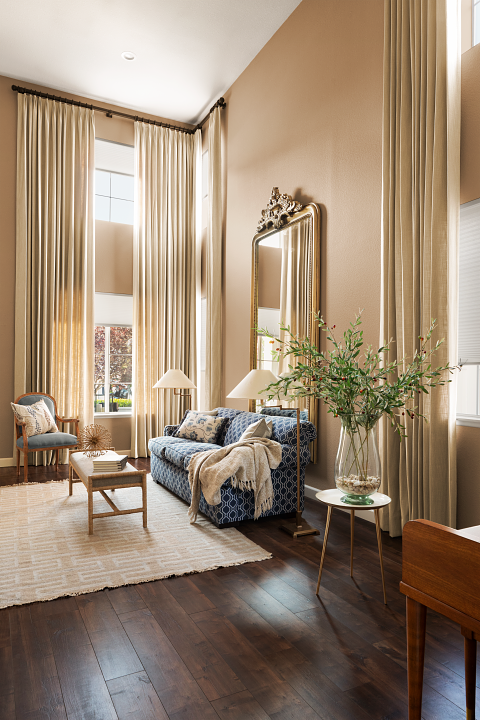Bright living room with tall beige curtains, a blue patterned sofa with pillows and a throw blanket, a glass vase with greenery on a small table, and a large mirror on the wall.