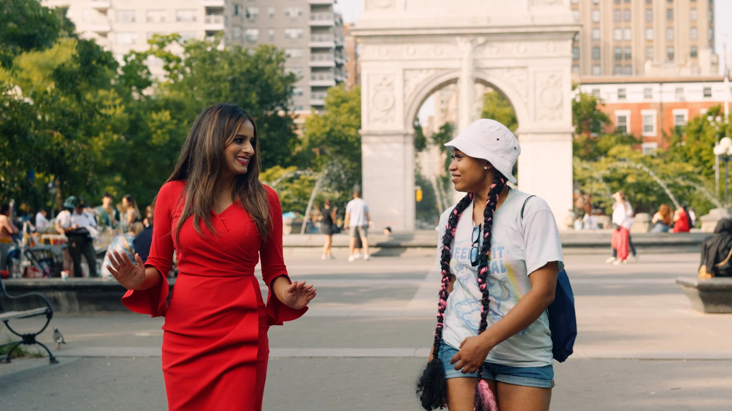 Two women talking and smiling in an urban park with the Washington Square Arch in the background. One woman is wearing a red dress, and the other is wearing a white t-shirt, denim shorts, and a white bucket hat.