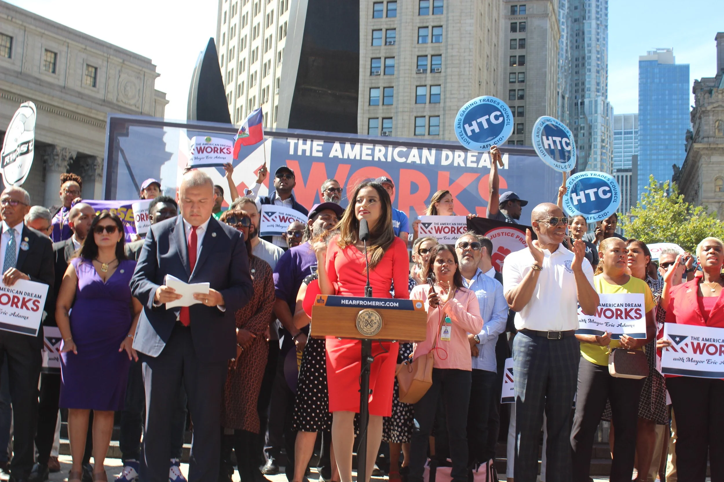 A crowd of people gathered outdoors at a protest or rally, some holding signs and banners that read 'The American Dream Works' and 'HTC,' with a woman speaking at a podium.