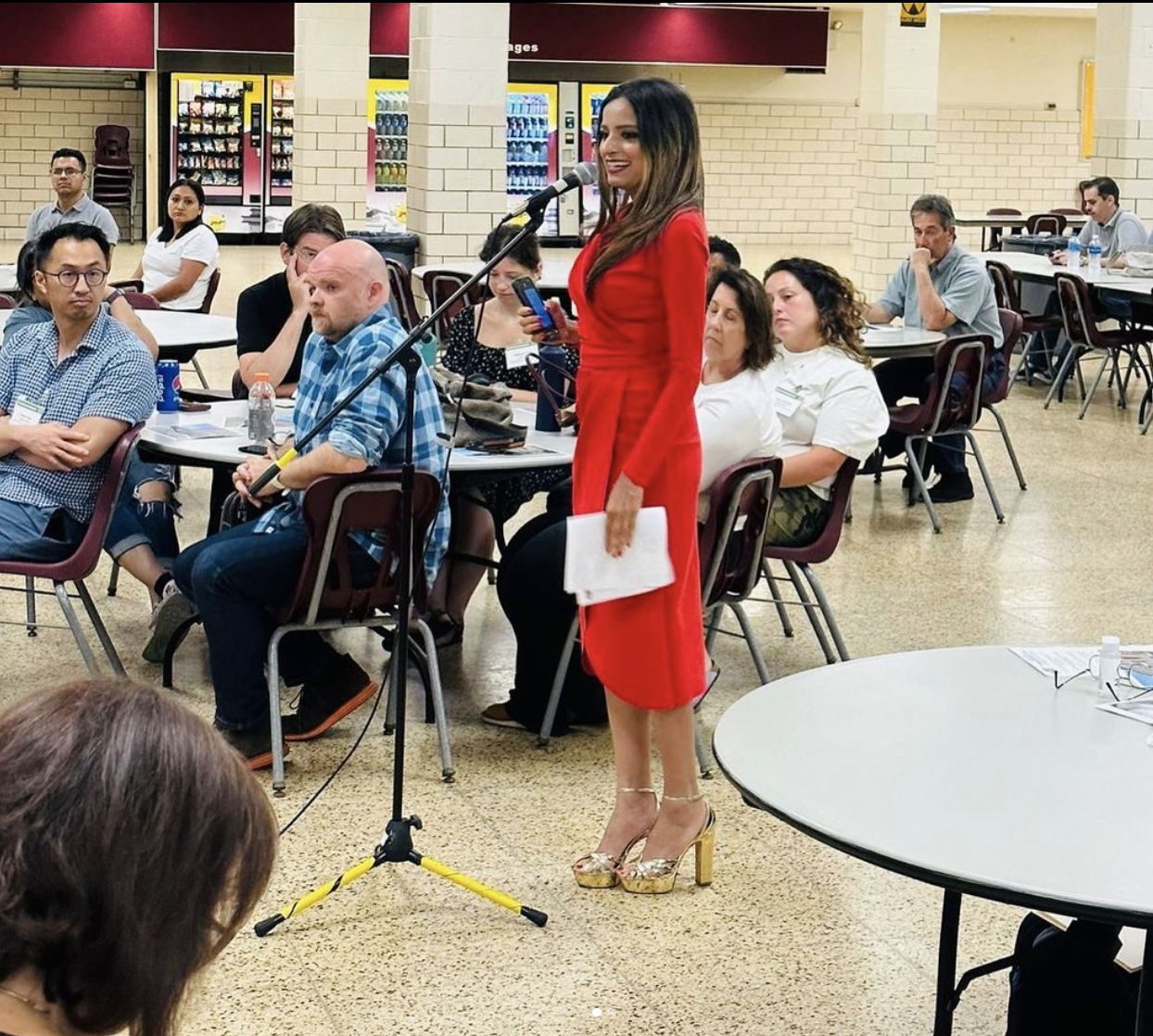 A woman in a red dress with high-heeled shoes is speaking into a microphone at a public event or meeting, holding a piece of paper in her hand, while a seated audience listens attentively.