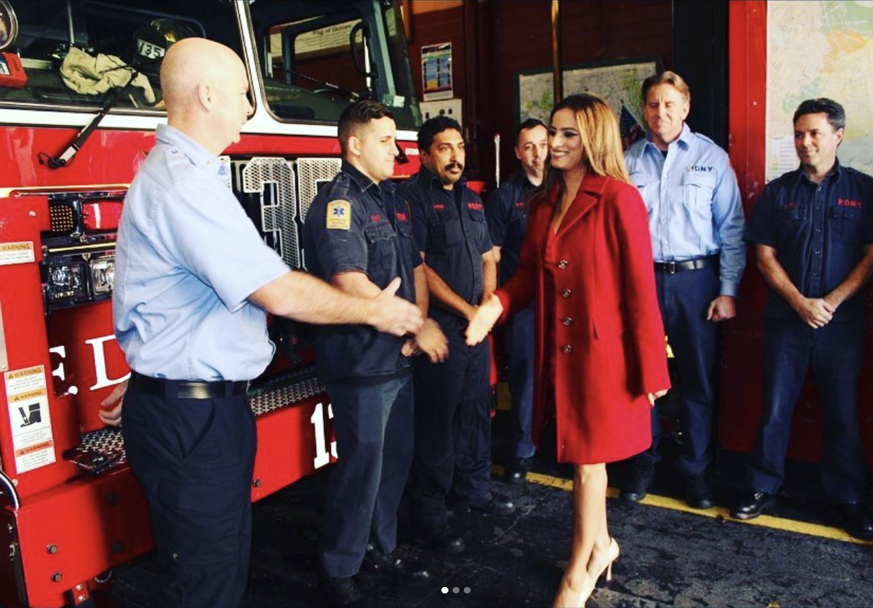 A woman in a red coat shaking hands with a firefighter in front of a fire truck, surrounded by other firefighters and coworkers.