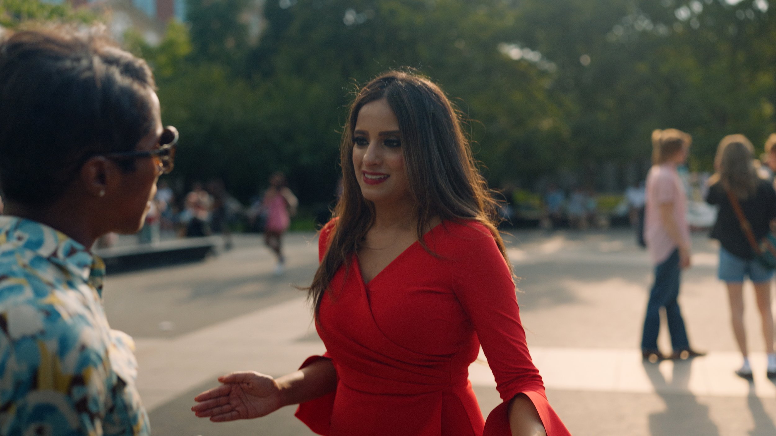 A woman in a red dress talking to an older woman with glasses outdoors in a park on a sunny day. Other people are seen in the background with trees.