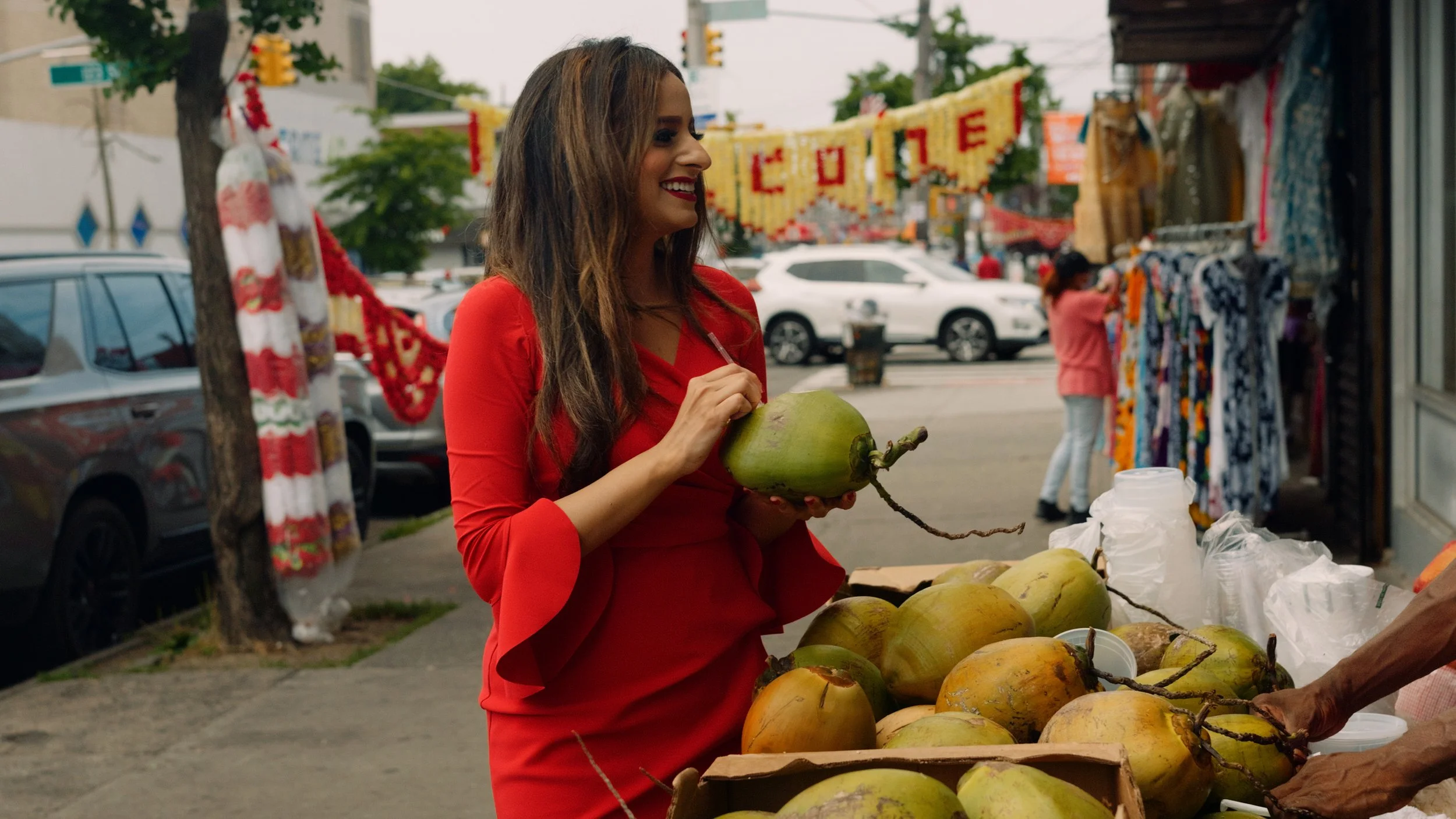 A woman in a red dress shopping for coconuts at an outdoor market, with vendors and cars in the background.