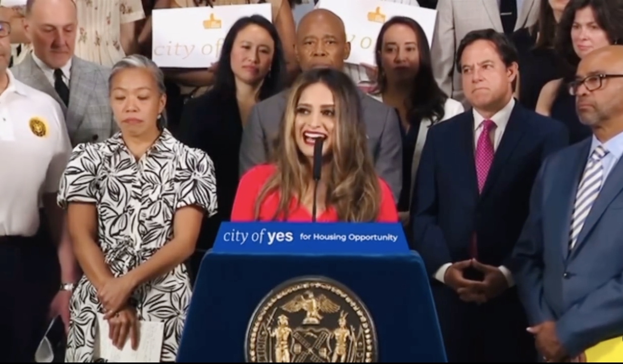 A woman at a podium speaking with a microphone, surrounded by a diverse group of people, including men and women, at a formal event about housing opportunities.