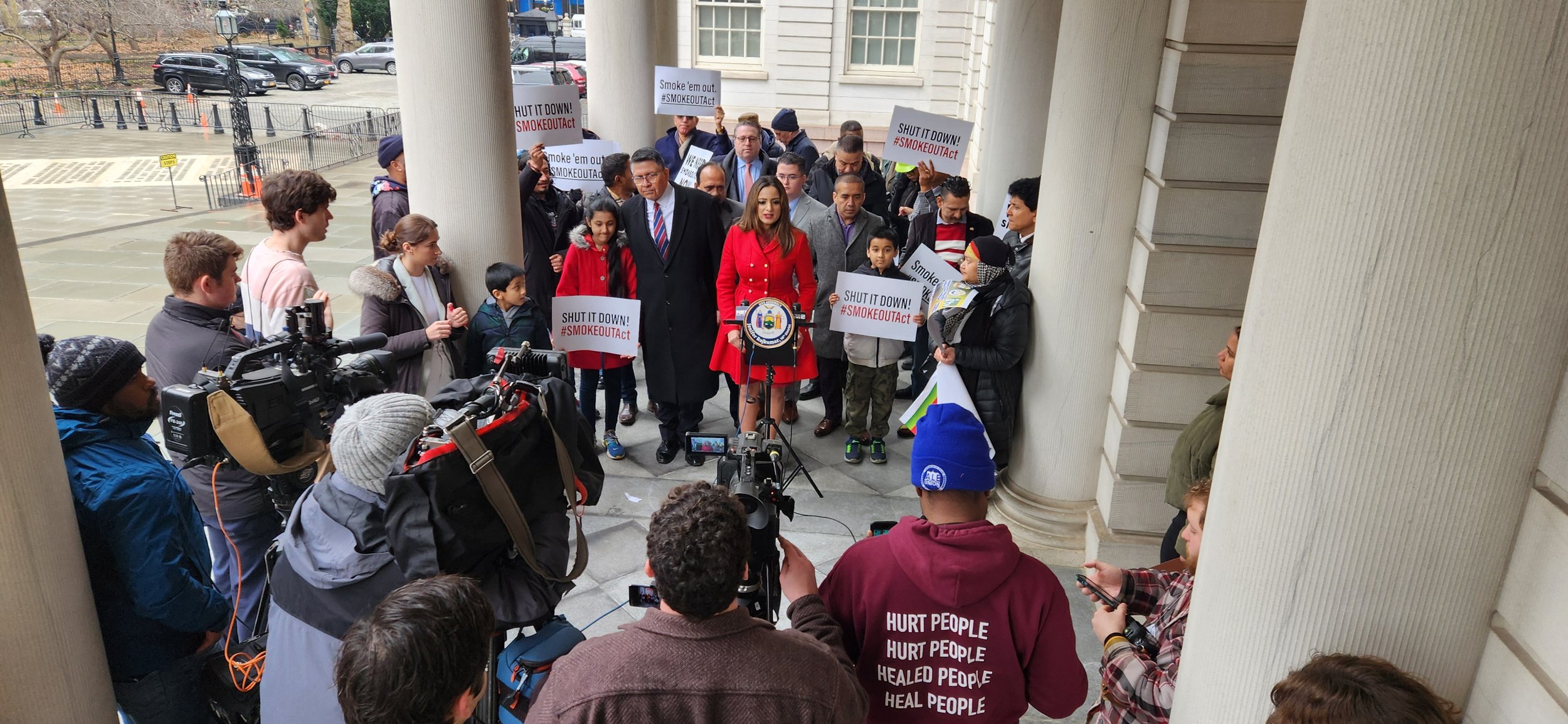 A group of people gathered on the steps of a building during a protest or rally, holding signs that say 'SHUT IT DOWN! #SMOKEOUTAct' and 'Smoke 'em out! #SMOKEOUTAct.' A woman in a red coat is speaking at a podium with a microphone. There are reporte
