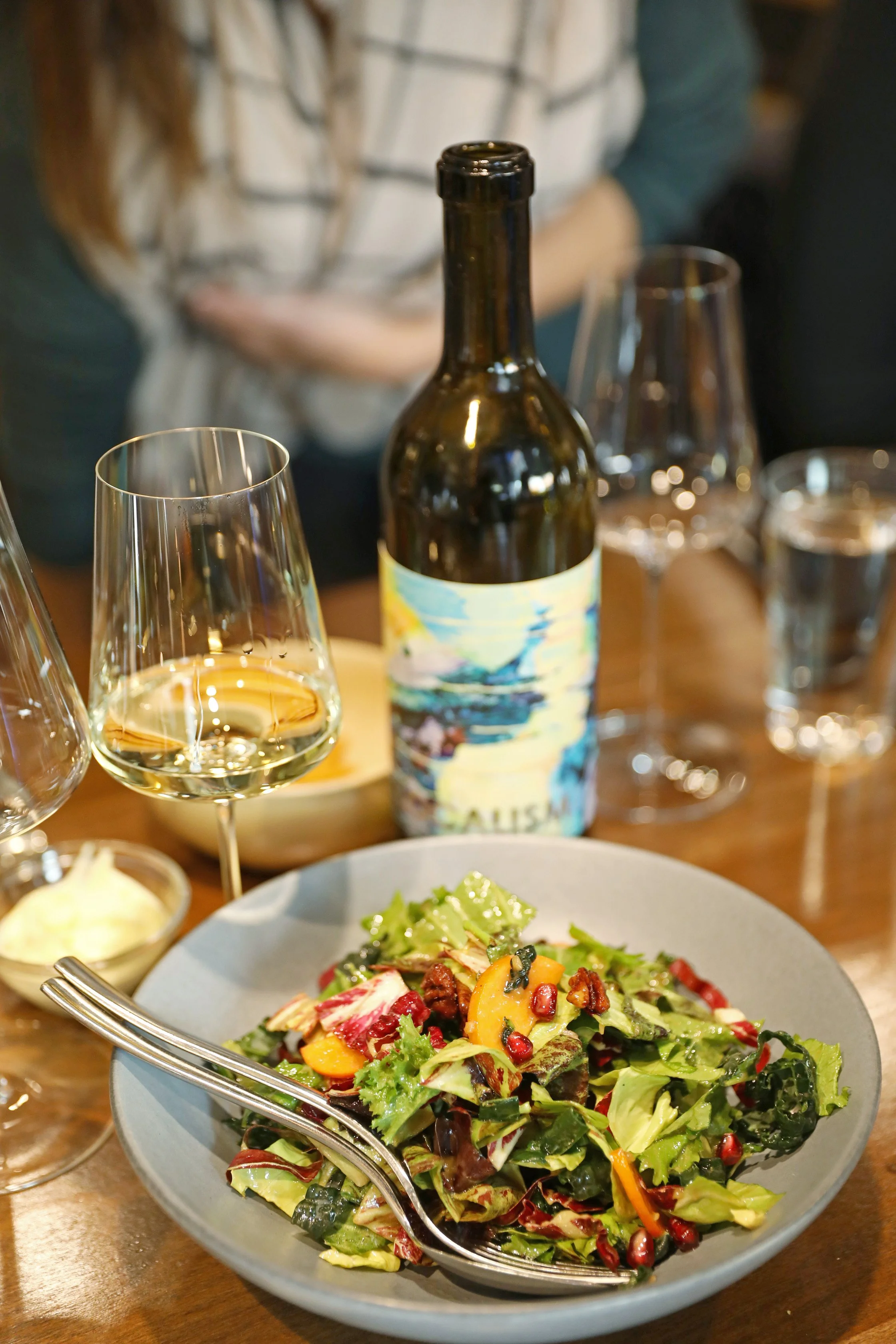A bowl of mixed greens salad with chopped vegetables and pomegranate seeds on a wooden table, with a fork resting in the bowl. In the background, there is a bottle of wine with a colorful label, and a glass of white wine alongside other wine glasses and a partially blurred person.