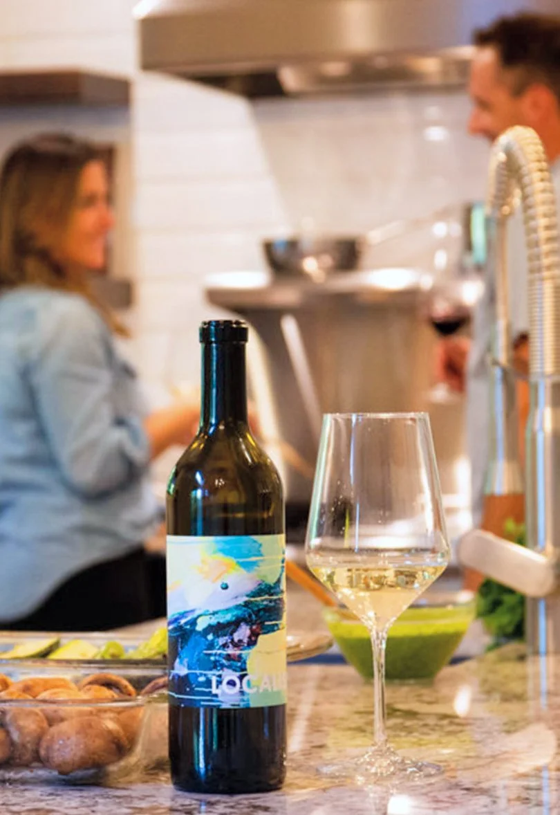 A bottle of white wine and a filled wine glass on a kitchen island, with two people talking and cooking in the background.