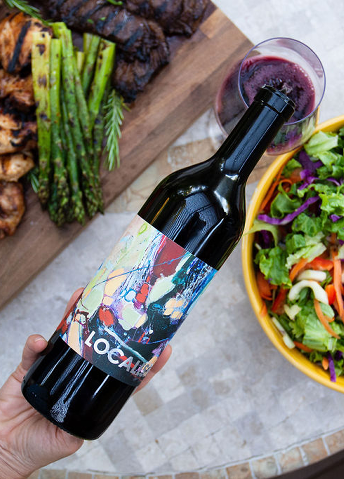 Person holding a wine bottle with an abstract colorful label, with a salad and grilled food on a wooden serving board in the background.