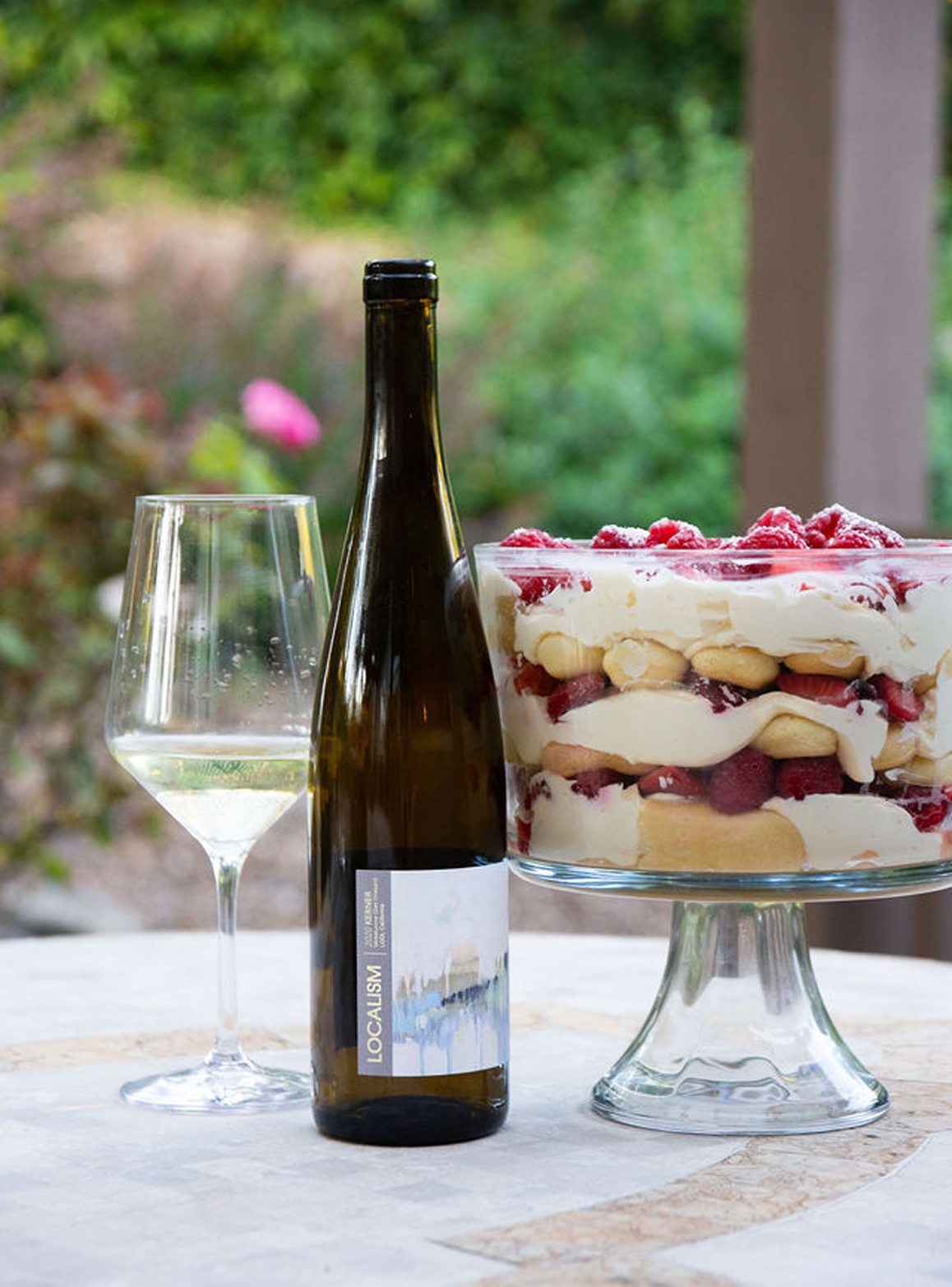 A bottle of white wine, a wine glass, and a layered strawberry and cream dessert on a table outdoors with greenery in the background.