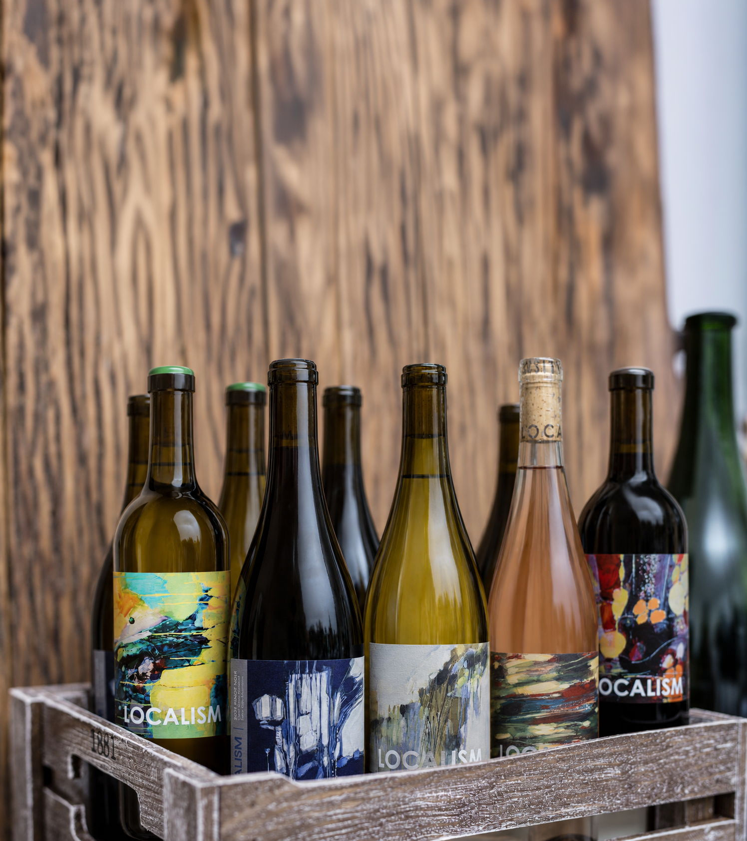 A wooden crate containing several bottles of wine with colorful, artistic labels, placed in front of a wooden background.