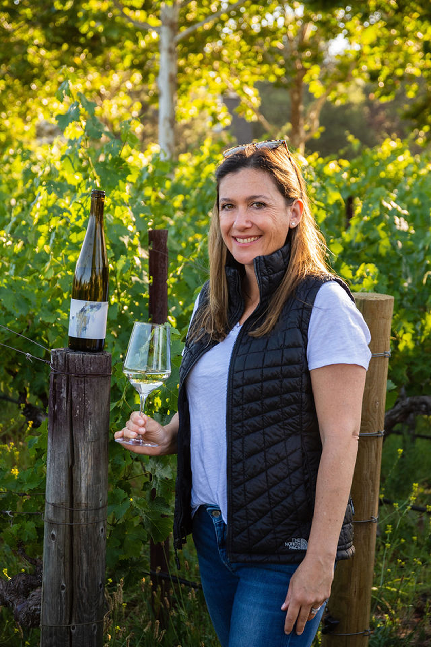 Woman in a vineyard holding a glass of white wine, standing next to a wine bottle on a wooden post, surrounded by lush green grapevines with sunlight filtering through the leaves.