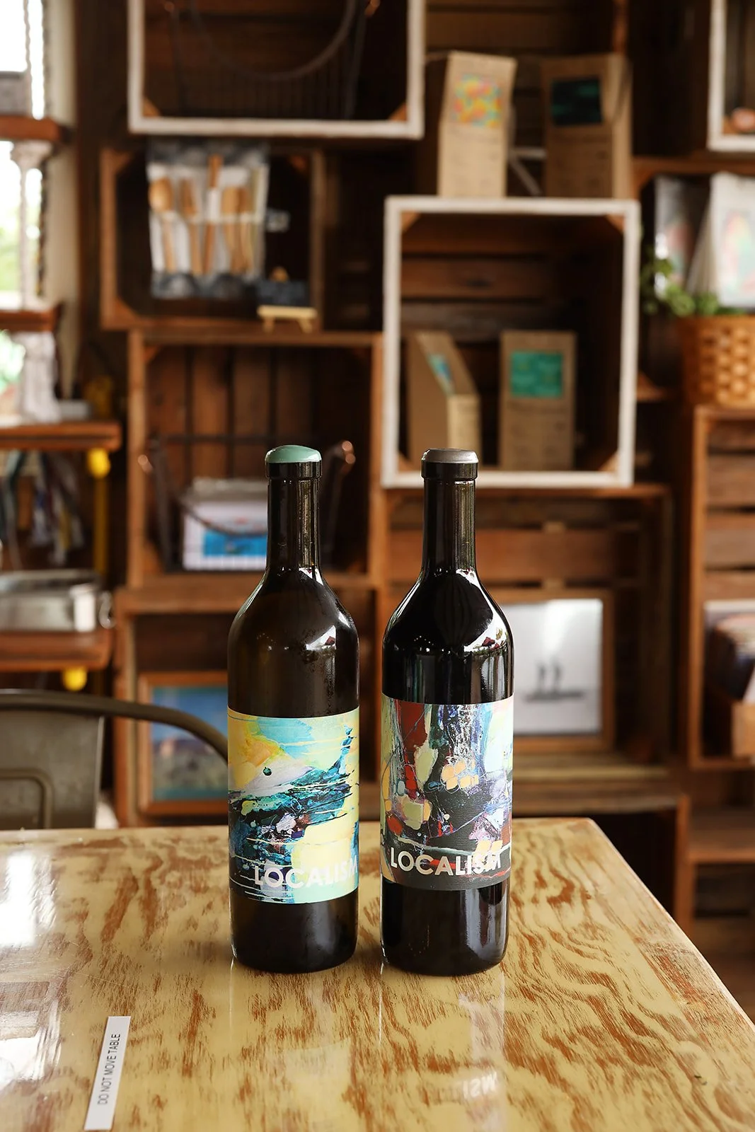 Two wine bottles with colorful abstract labels placed on a wooden table in a cozy, rustic setting with wooden display shelves in the background.