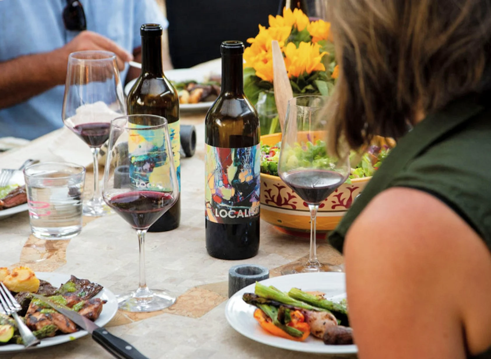 A table set for a meal with two bottles of wine, several glasses of red wine, a salad, and plates of food. There are flowers in a yellow and red bowl in the background. People are sitting around the table.