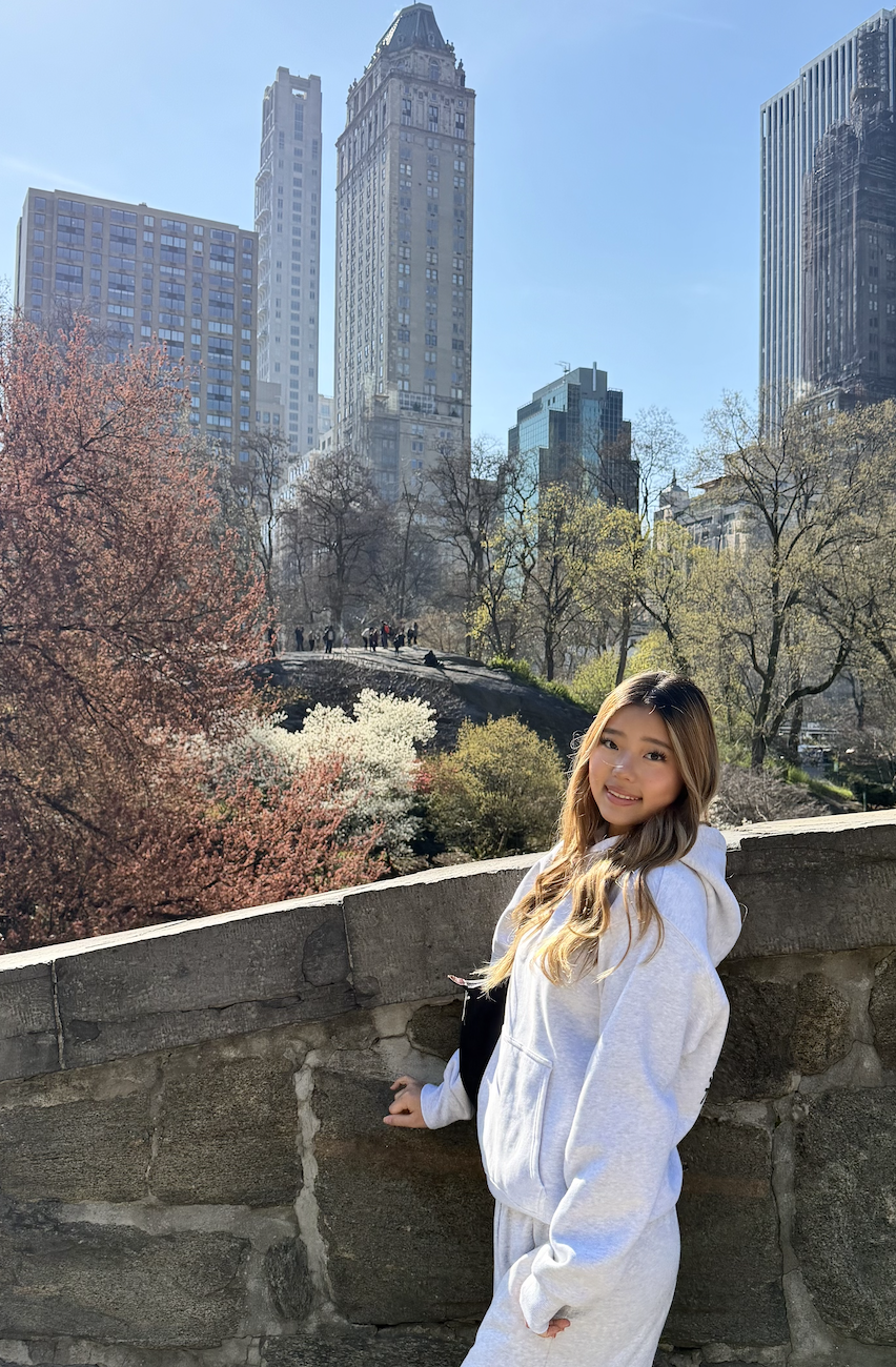 Young woman with long, wavy hair smiling and standing near a stone wall in Central Park, New York City, during spring with blooming trees and tall skyscrapers in the background.