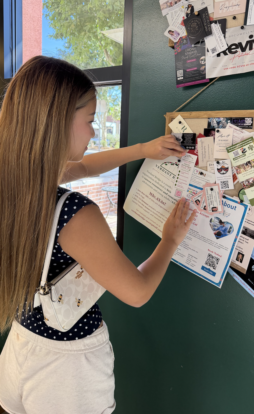 Young girl reading a poster and looking at business cards and flyers on a bulletin board inside a building near a window with a brick building and trees outside.