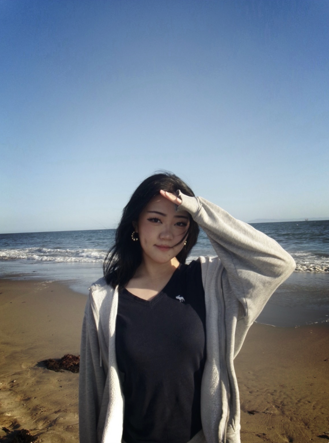 A young woman standing on a sandy beach with ocean waves behind her, saluting with her right hand while shielding her eyes from the sun.