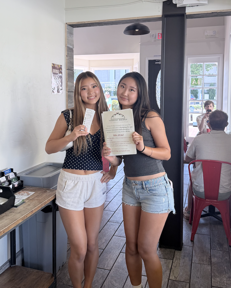 Two young women standing inside a restaurant, smiling and holding flyers or brochures. One has long blonde hair and is wearing a black polka dot top with white shorts, the other has long dark hair and is wearing a gray crop top with denim shorts.