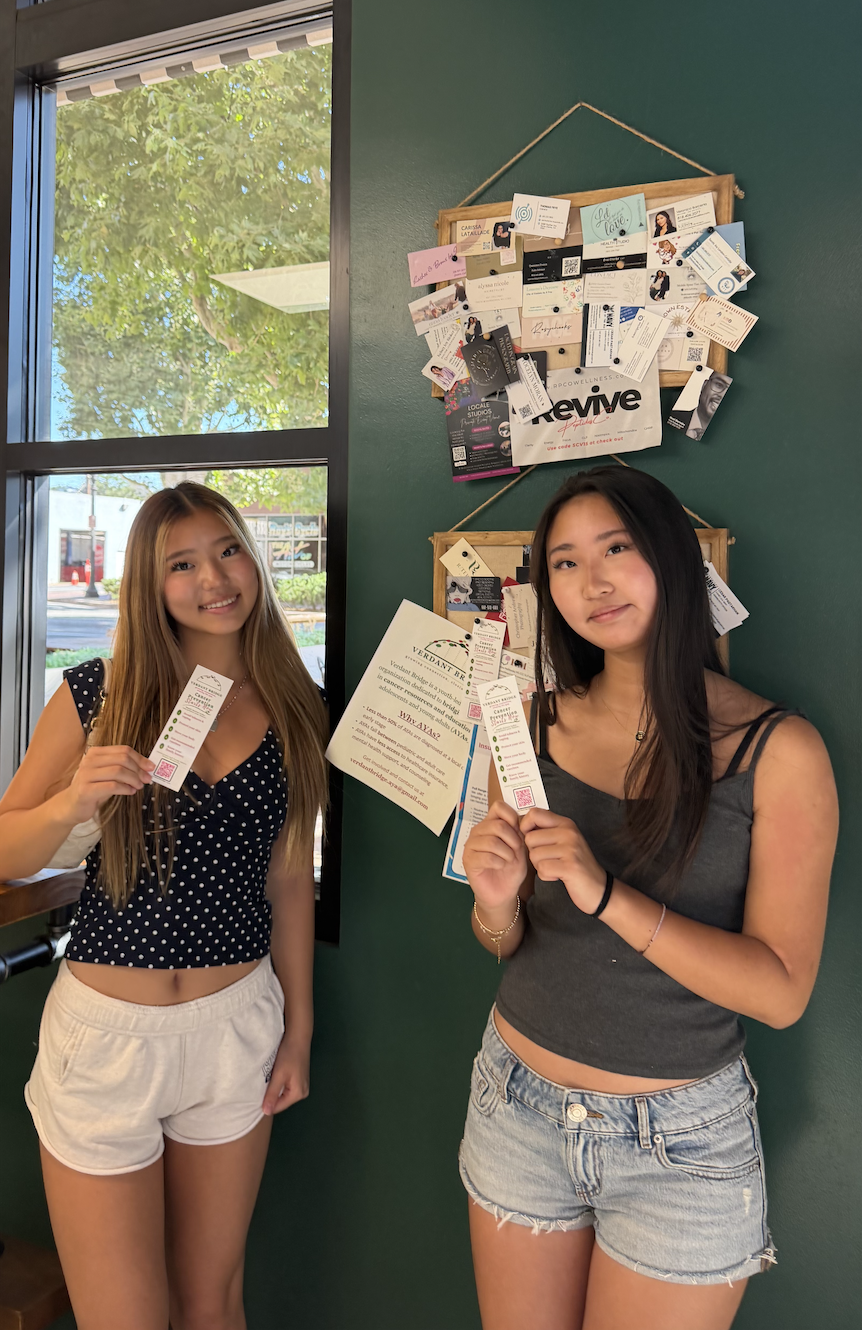 Two young women standing inside a room near a window holding what appear to be business cards, with a green wall behind them decorated with a bulletin board filled with various flyers, notes, and cards. Outside the window, trees and a street scene are visible.