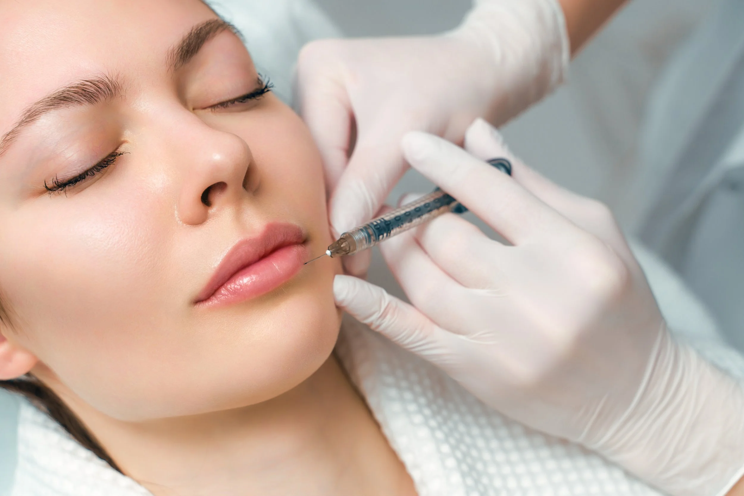 A woman with closed eyes receiving a cosmetic injection in her cheek from a medical professional wearing gloves.