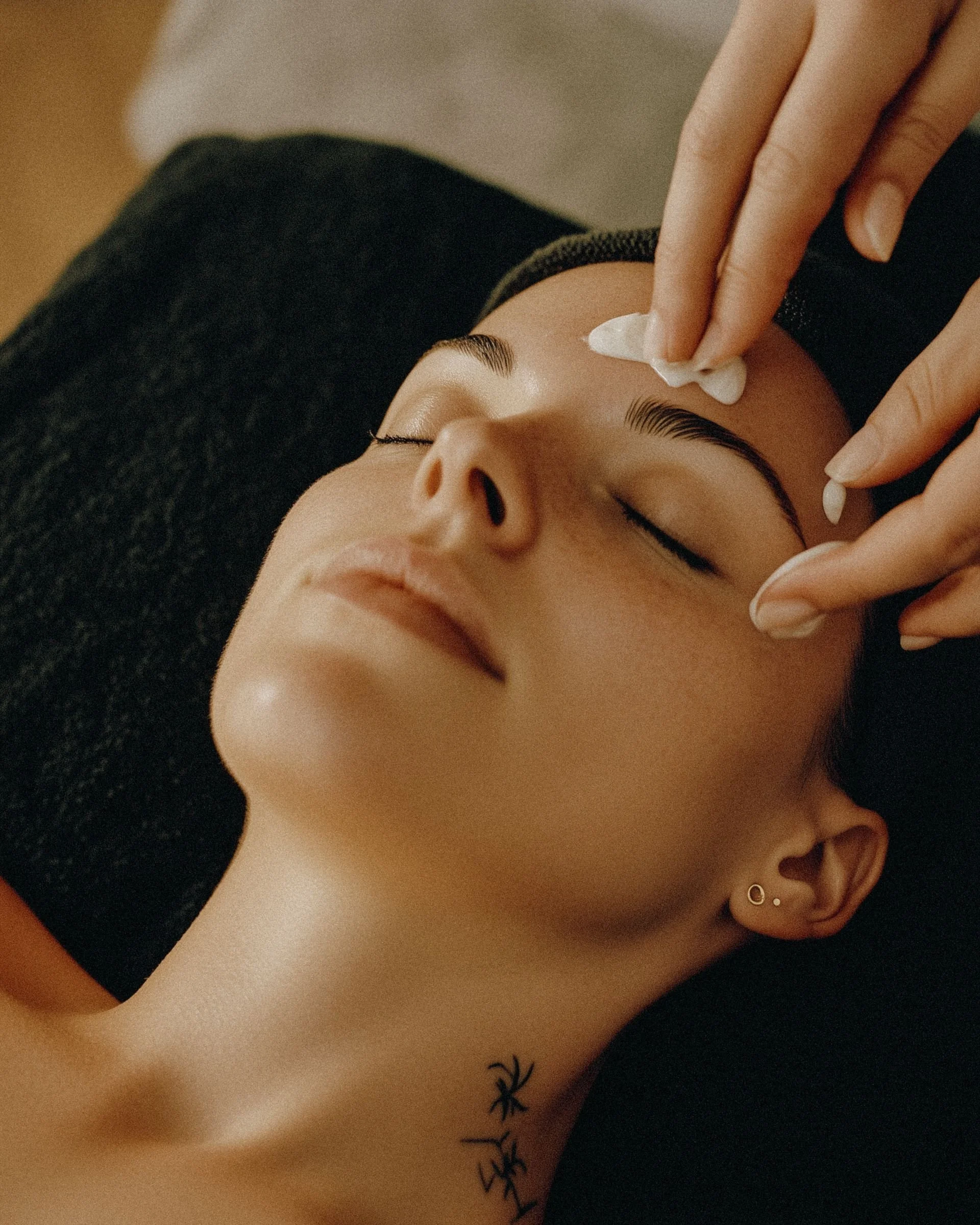 Close-up of a woman lying down with her eyes closed, receiving a facial treatment, with a cotton pad on her forehead and wearing earrings, black sweater, and showing a small tattoo on her chest.