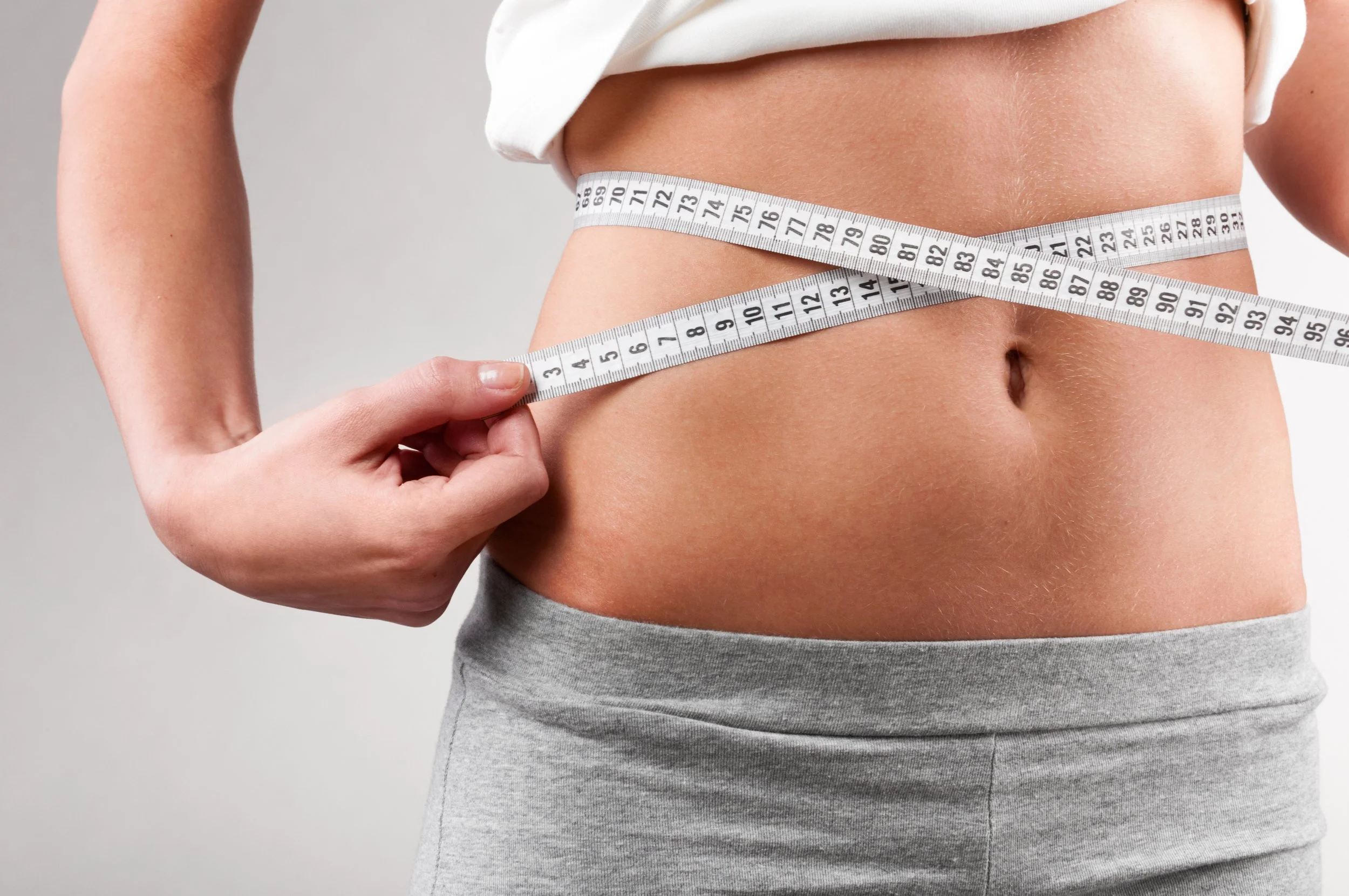 A person measuring their waist with a tape measure, wearing a white top and grey sweatpants.