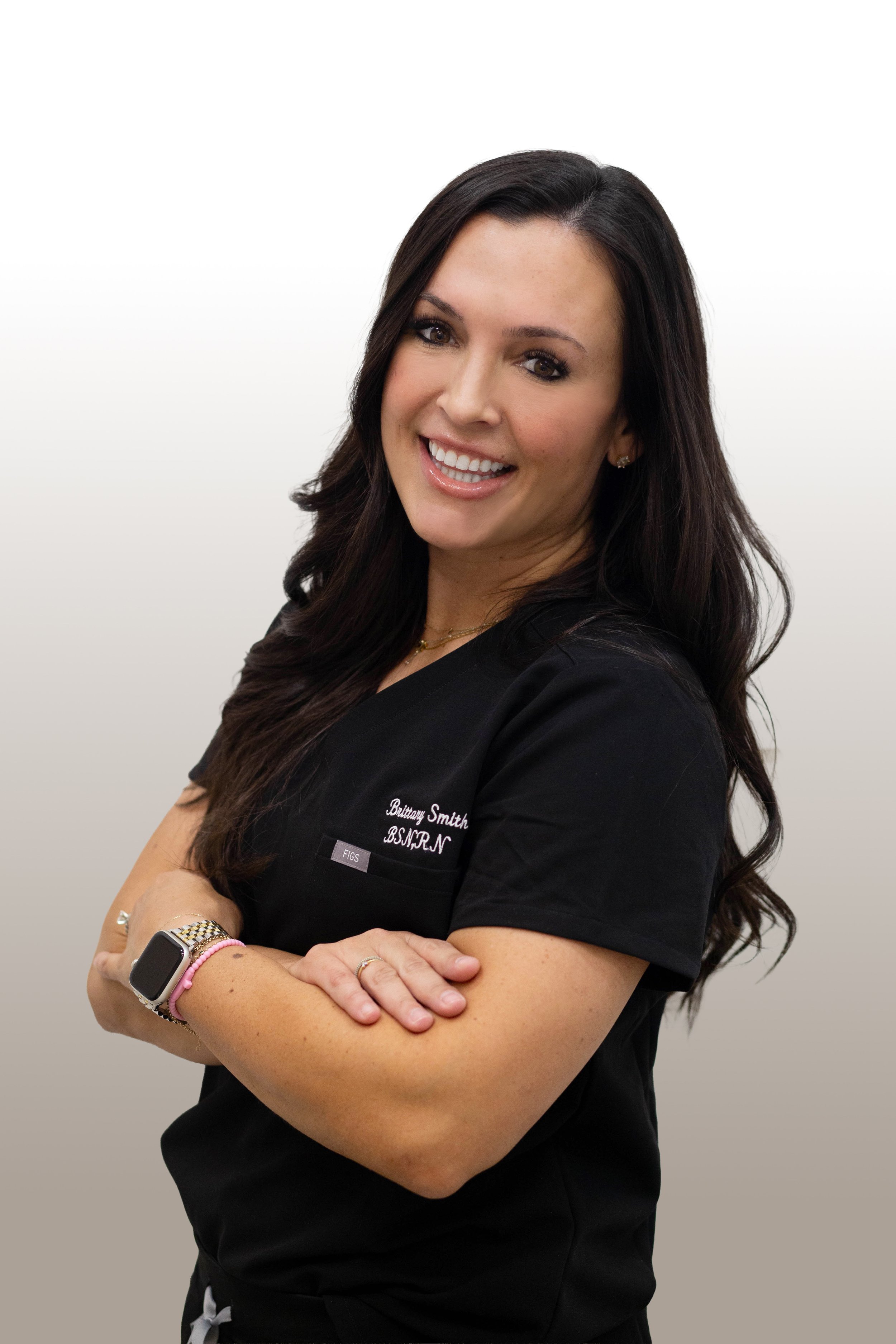 A woman with long dark hair smiling with arms crossed, wearing a black medical scrub top with a name tag that reads Brittany Smith, B.S., N.P., and a smartwatch on her left wrist.