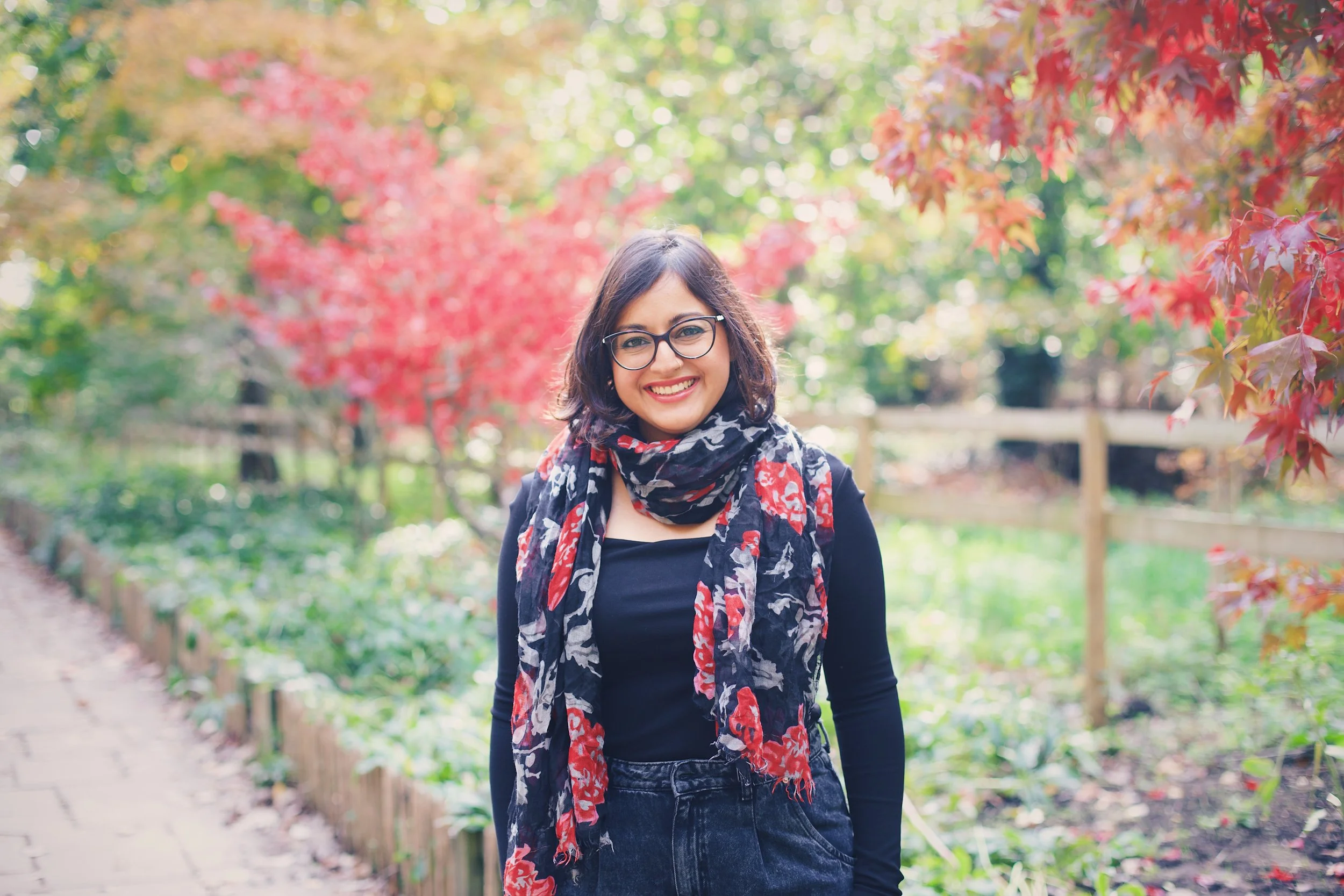 A woman with glasses, dark hair, wearing a black top and a colorful scarf, smiling outdoors in a park with autumn foliage.