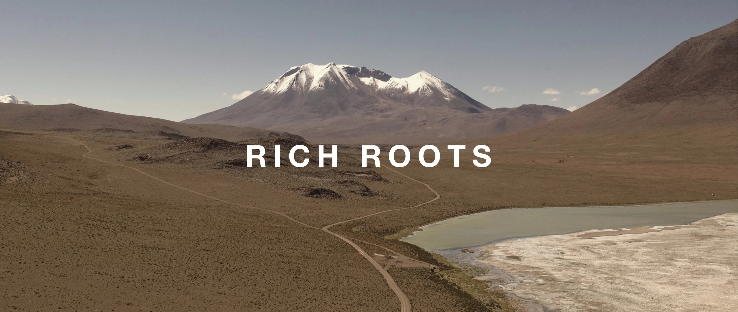 A mountain with snow on its peak in a desert landscape with a winding path and a body of water in the foreground, text overlays reading 'Rich Roots'.