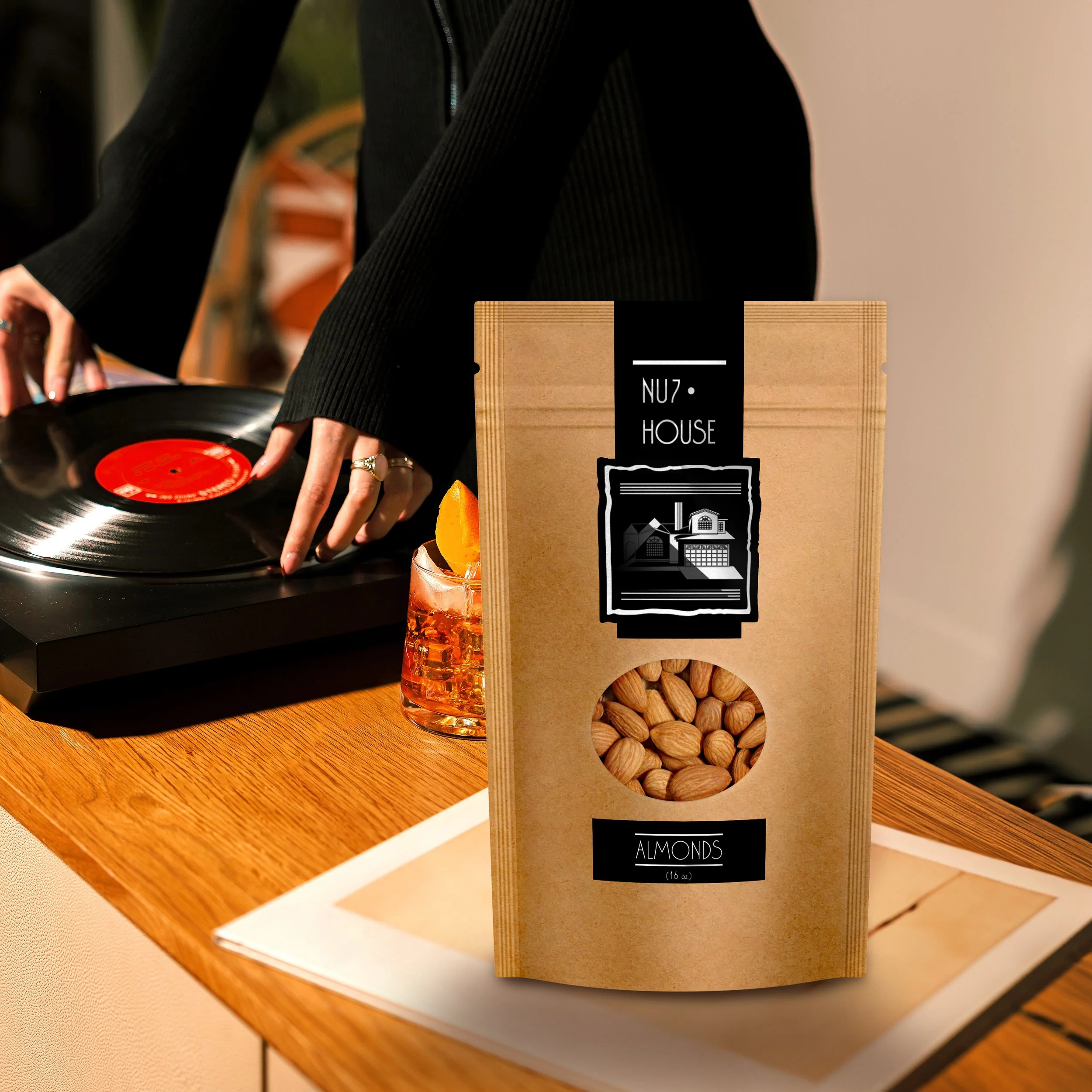 A person with a black long sleeve top using a turntable with a vinyl record. There is a brown paper bag labeled 'Almonds 16 oz.' on the table next to the turntable.