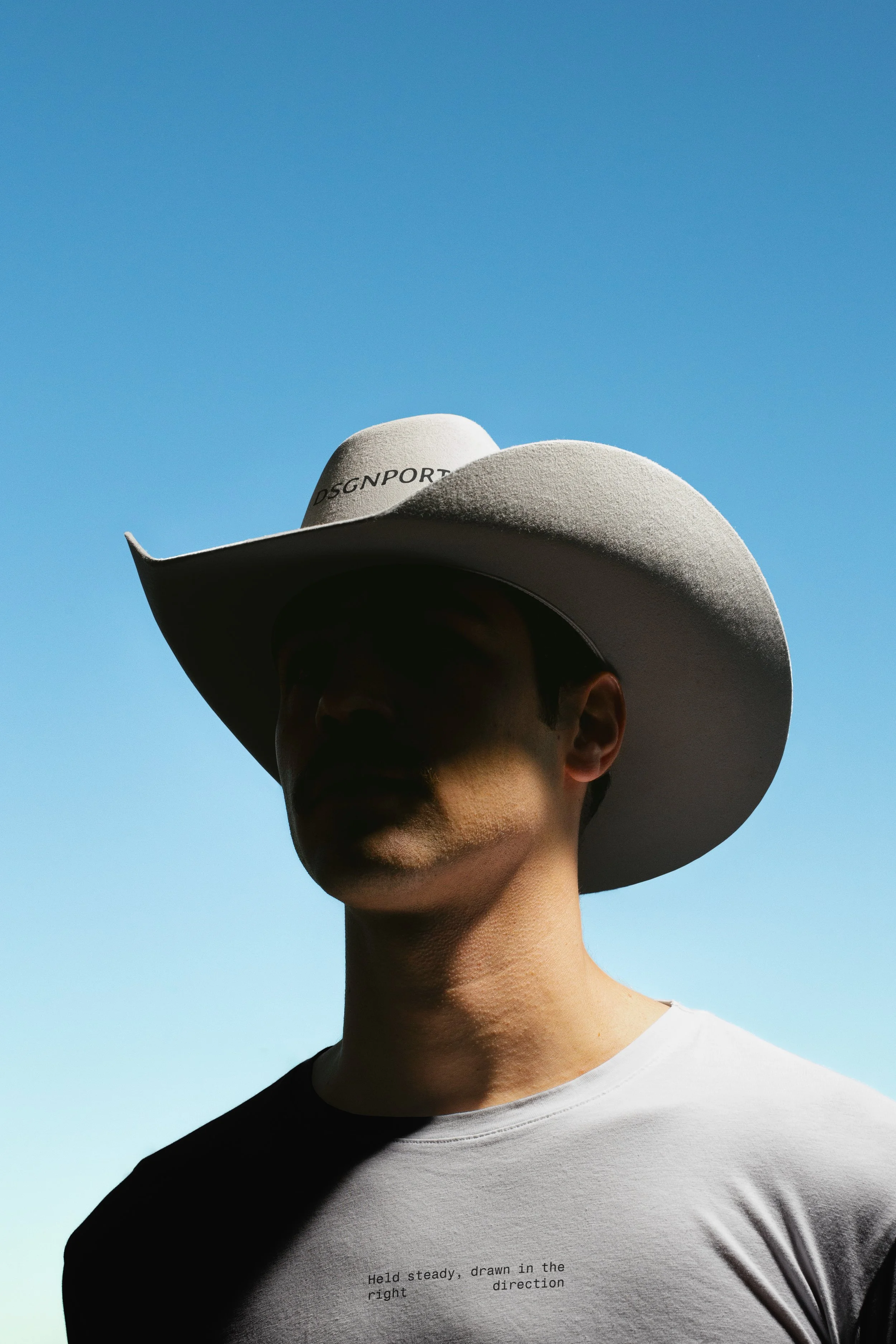 A person wearing a white wide-brimmed hat and a white T-shirt with text, standing outdoors against a clear blue sky. The person's face is in shadow, and their head is slightly turned to the side.