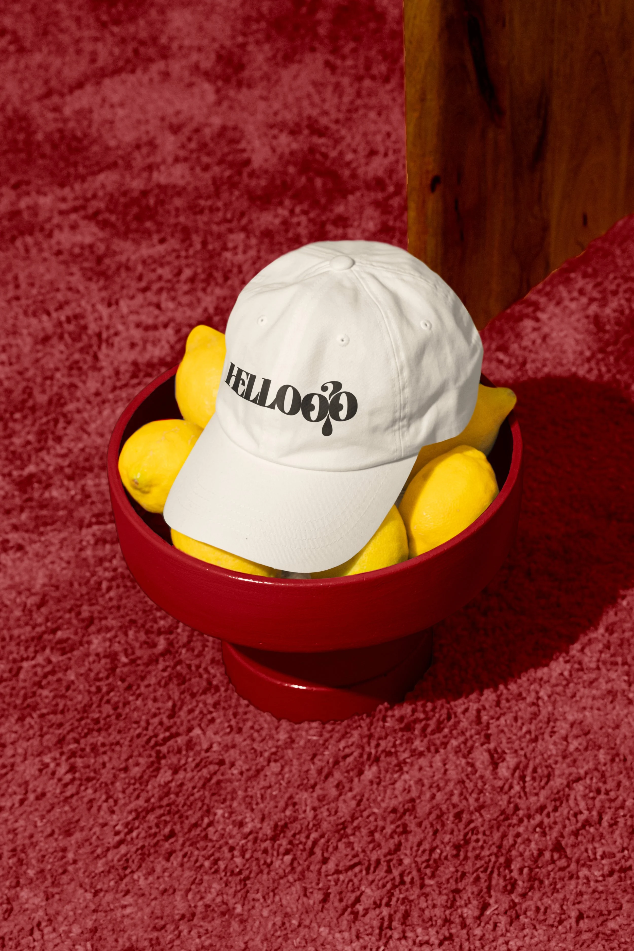 A white baseball cap with black text resting on several yellow lemons in a red bowl, placed on a textured red carpet floor next to a wooden surface.
