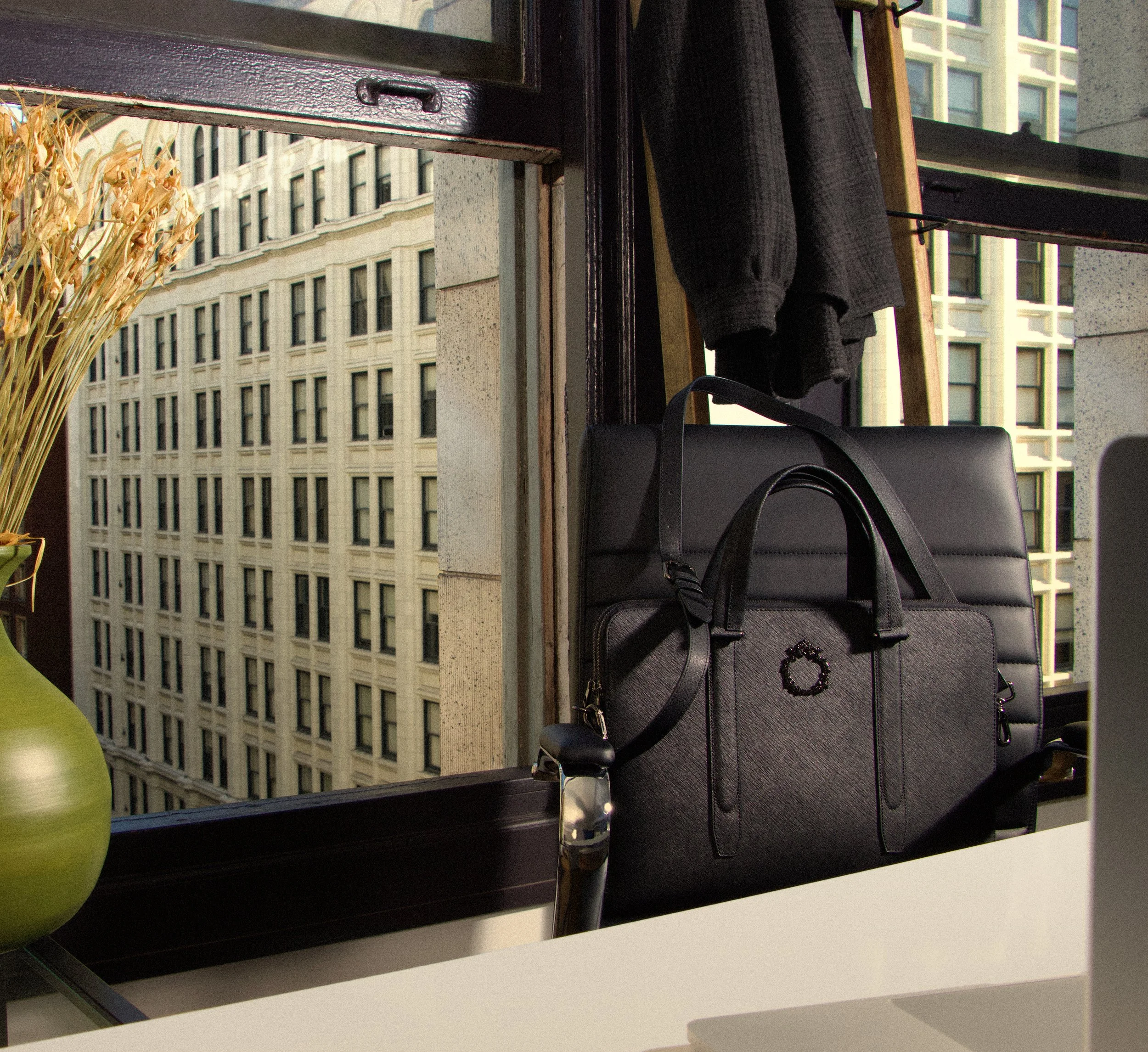 A desk view showing a green vase on the left, a black handbag with a metal pomegranate logo on the front, a black chair, black coat and a ladder behind, with large building windows visible outside.
