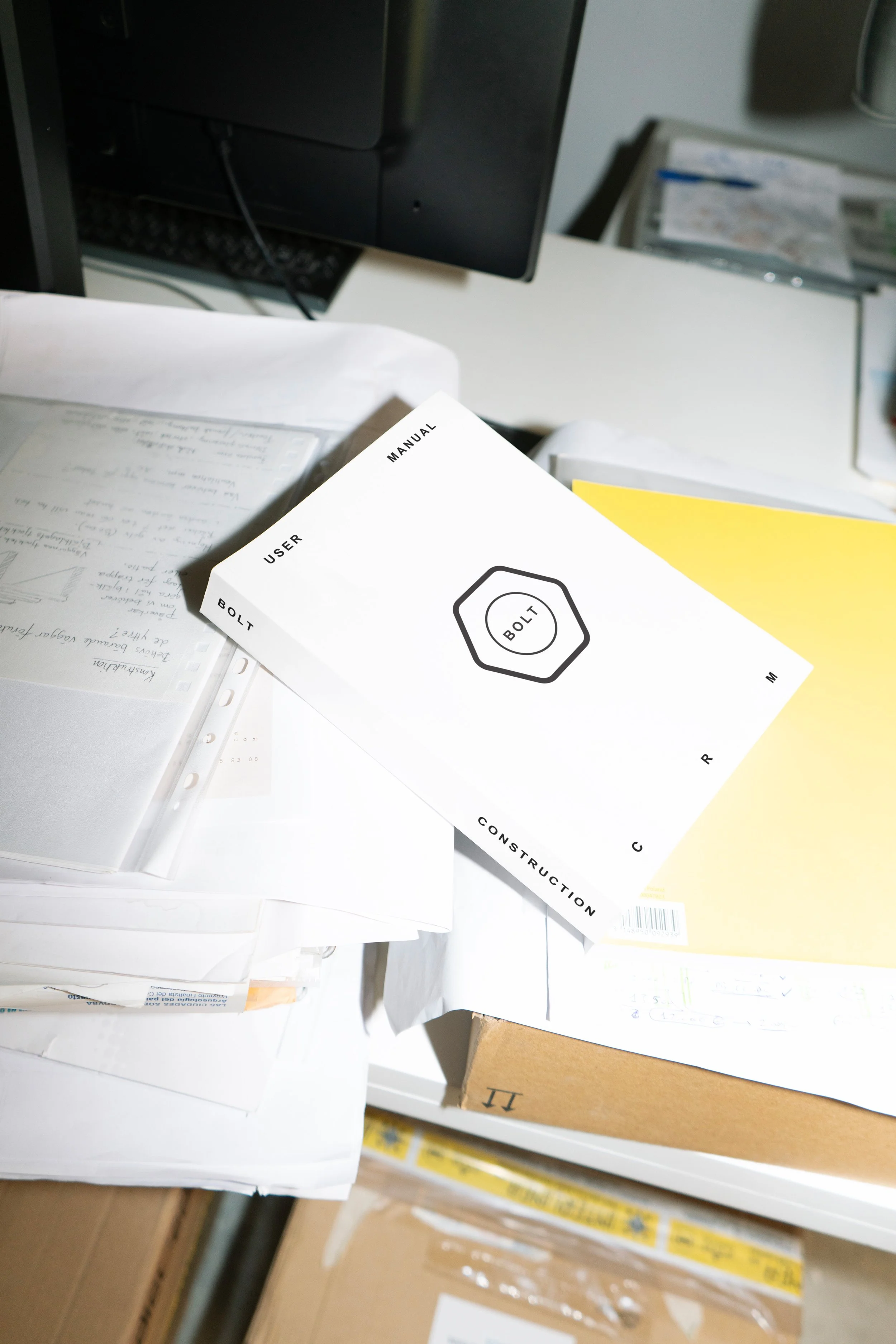 A cluttered desk with various papers, manuals, and boxes, including a white manual labeled 'BOLT' and 'USER MANUAL' on top of the papers.