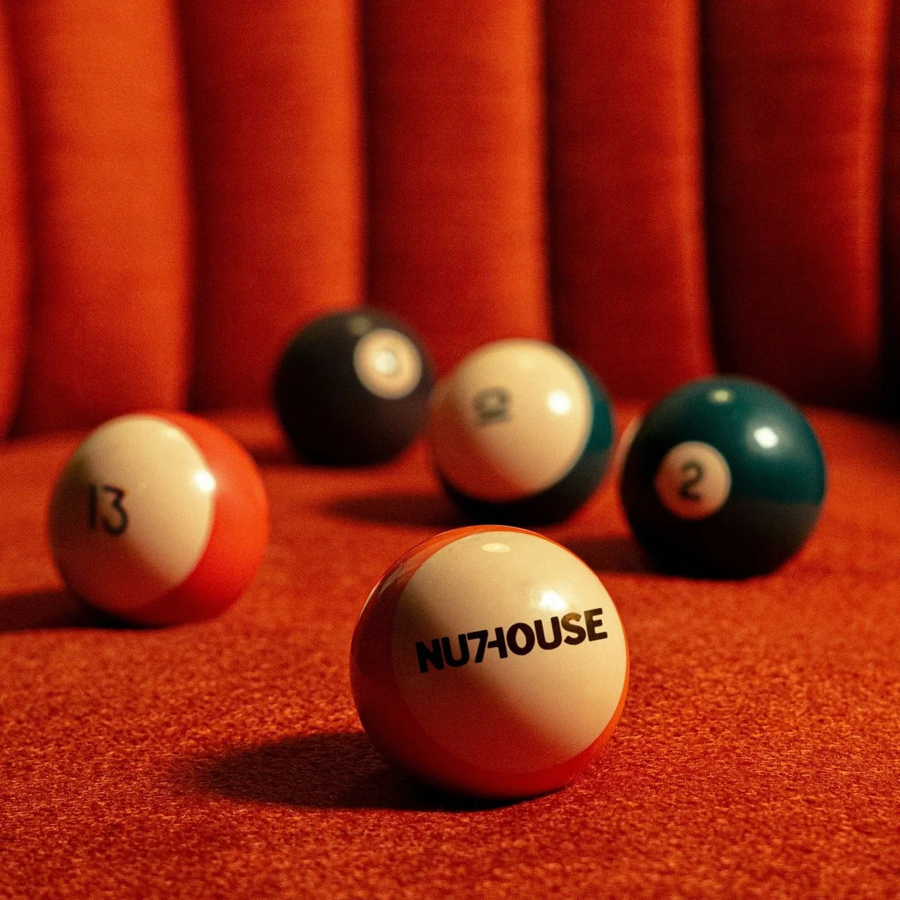 Six billiard balls arranged on a red billiard table, with a red curtain backdrop. The balls include the 13, 8, 2, and 12 balls, with the 8-ball in the background and the 13-ball in the foreground displaying the logo 'NU7HOUSE'.
