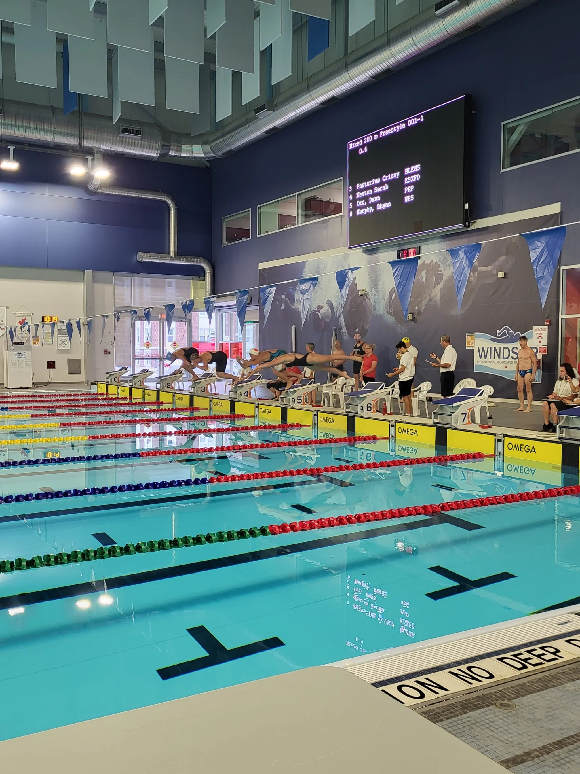An indoor swimming pool with swimmers on starting blocks, countdown clock, digital scoreboard, blue walls with large windows, and blue buntings hanging from the ceiling. The pool is divided by red, yellow, and blue lane dividers. There are people standing and sitting near the poolside and a large digital screen displaying swimmer names and event information.