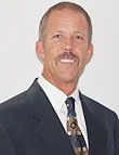 A middle-aged man with a mustache, wearing a dark suit, white shirt, and patterned tie, smiling.