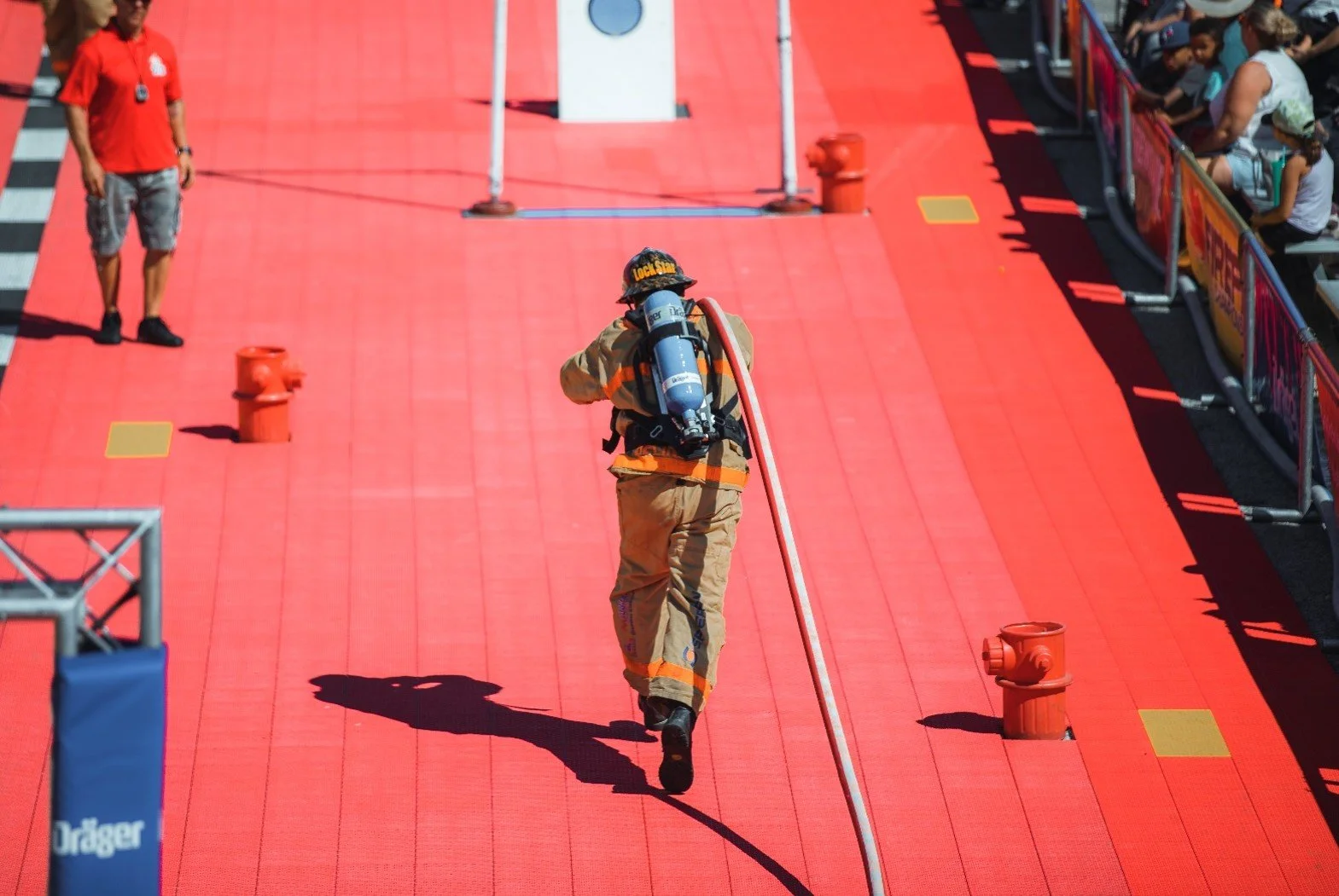 Firefighter walking on a red stage with a safety hose on their back, with spectators watching from the side.