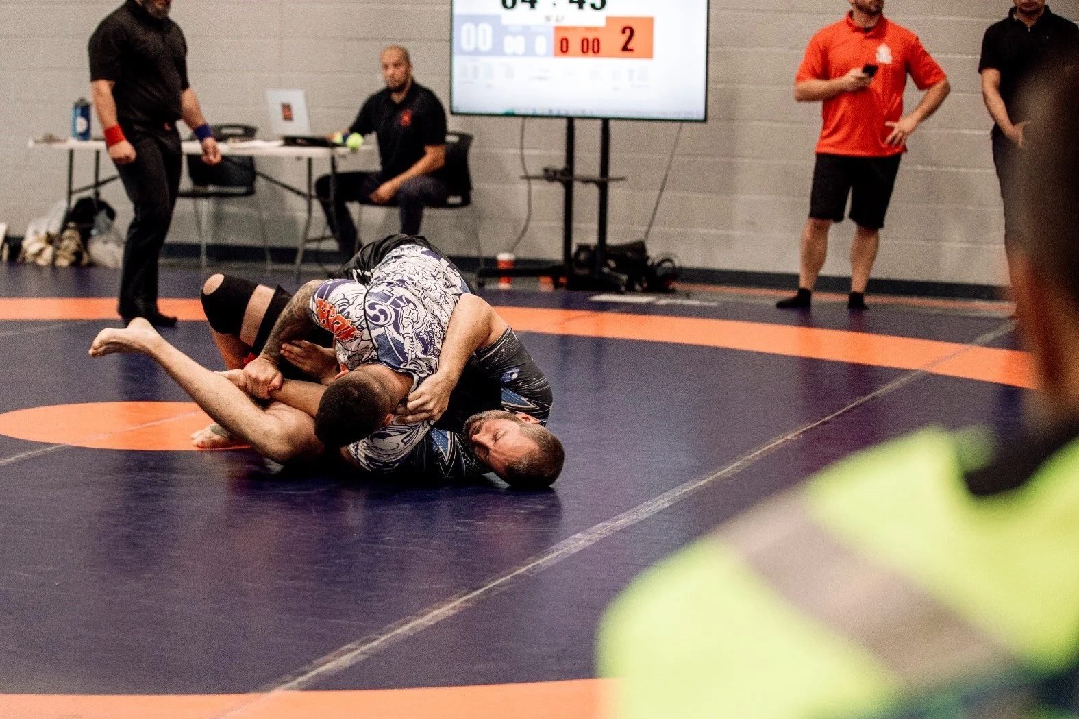 Two men competing in a grappling martial arts match on a blue and orange mat, with one applying a submission hold on the other. In the background, referees and spectators observe, along with a digital scoreboard displaying a close score.