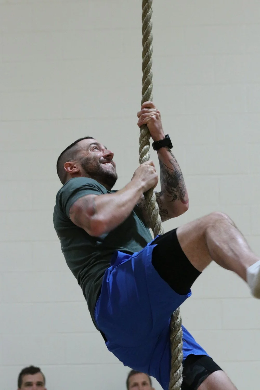 Man climbing a thick rope in a gym or indoor training facility with a beige wall in the background.
