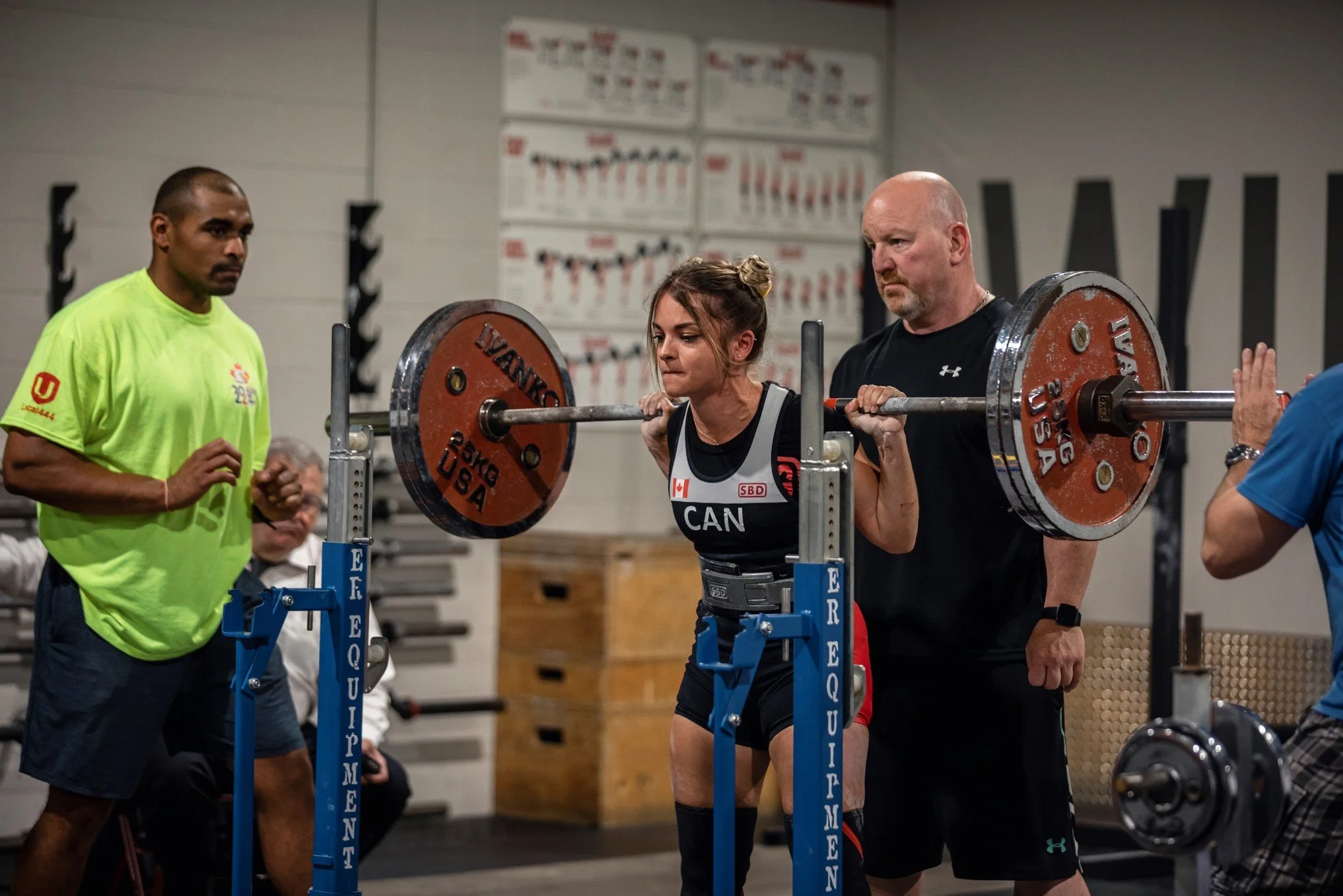 A woman performing a squat exercise with a barbell at a gym, assisted by two spotters, with one spotter on each side. The woman is focused and wearing a black workout outfit.