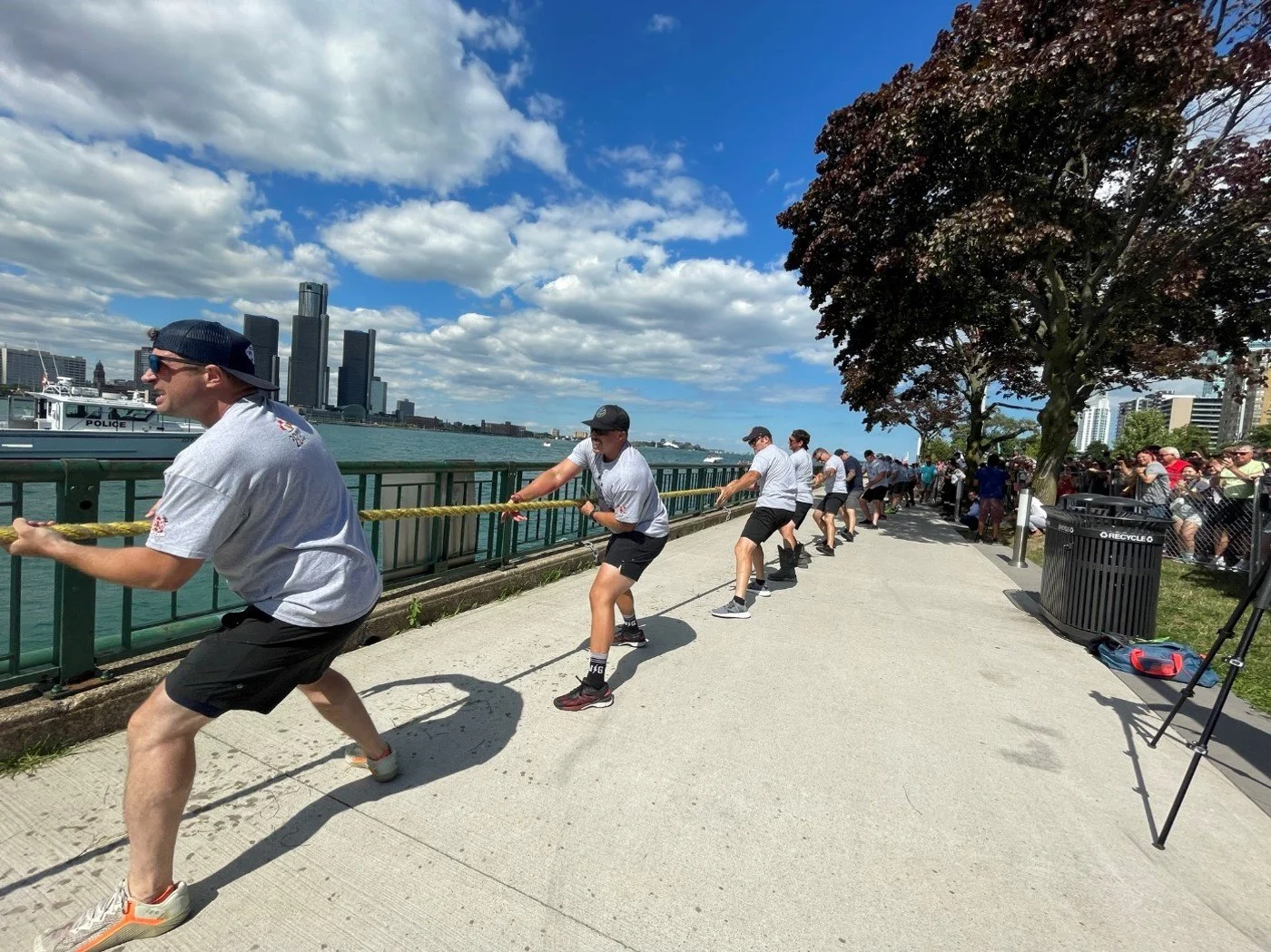 People participating in a tug-of-war game along a waterfront on a sunny day, with city skyline in the background and spectators watching.