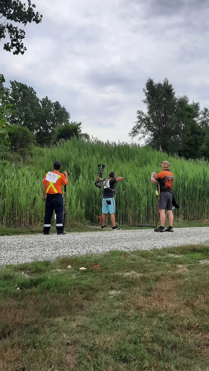 Three men are standing on a gravel path near tall green grass and trees, with one holding a bow and arrow and the other two speaking or observing. The sky is cloudy.
